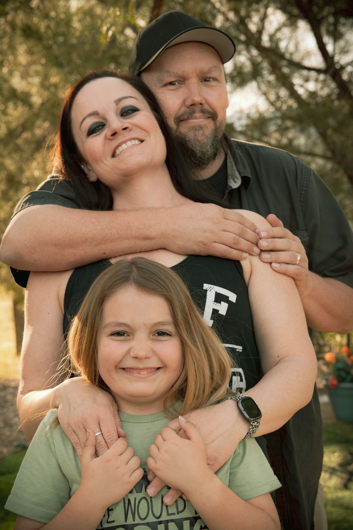 A smiling family of three poses outdoors, with parents embracing their young daughter among green trees.
