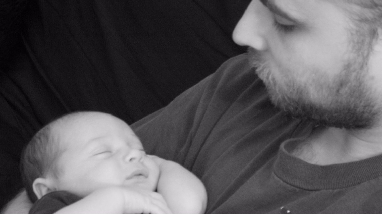 Black and white photo of a bearded man tenderly cradling a sleeping newborn baby in his arms.