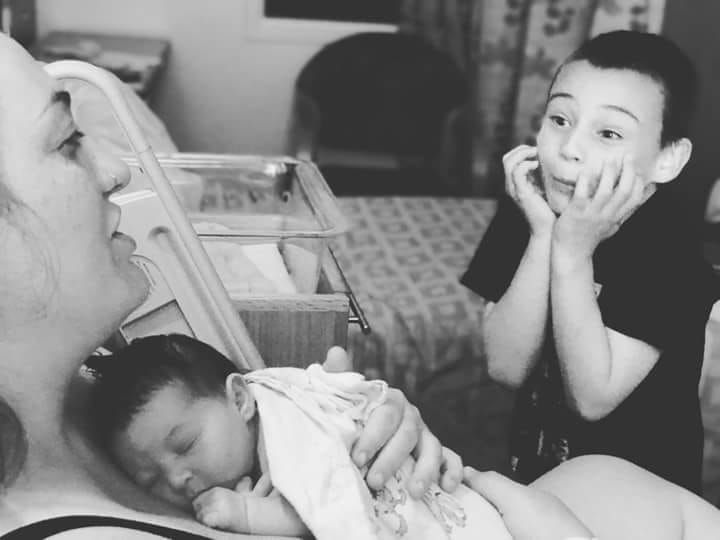 Black and white photo of a woman holding a newborn baby while an older child watches with amazement in a hospital room.