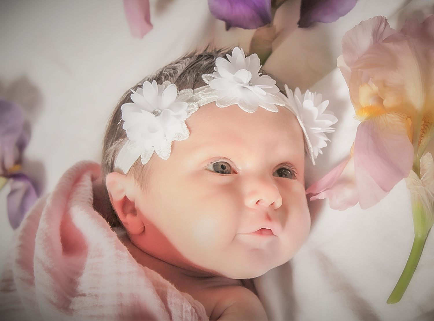Newborn baby girl wearing a white floral headband, wrapped in pink, surrounded by colorful tulips in a soft portrait.