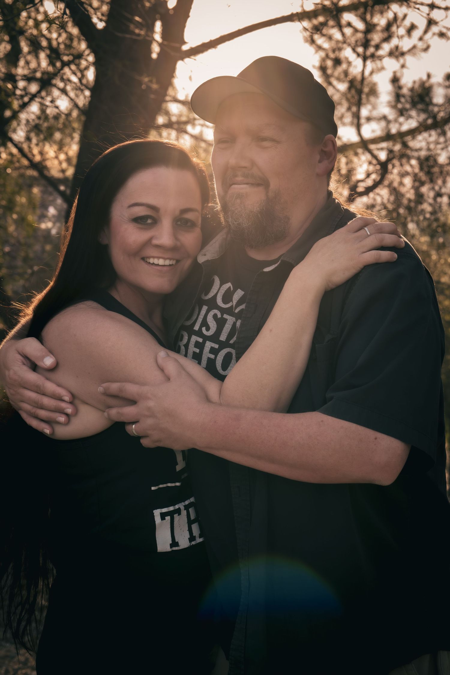 Couple embracing outdoors at golden hour, smiling woman with dark hair and bearded man in black caps, trees in background.