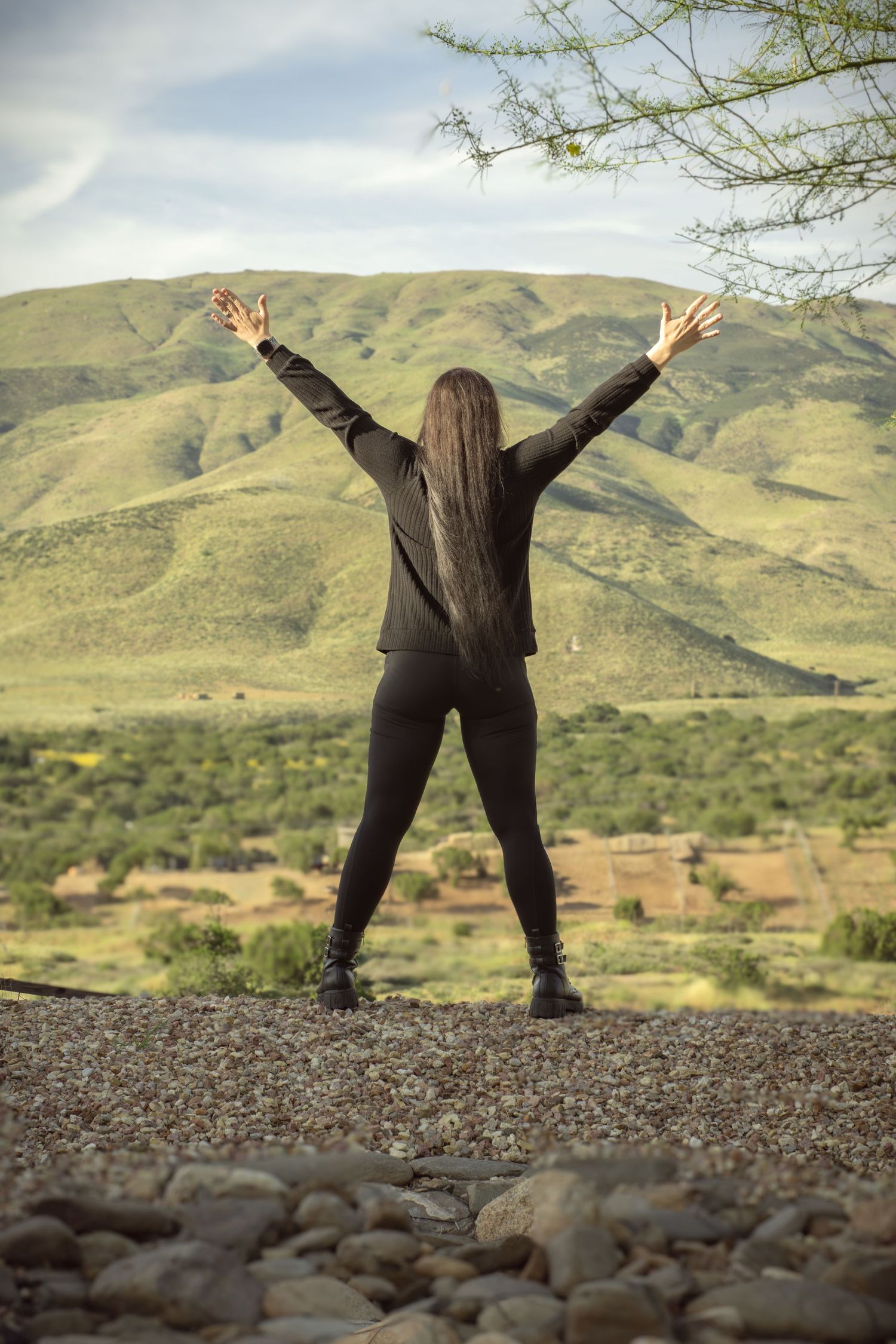 Woman in black outfit stands with arms raised facing lush green mountains and valley landscape.