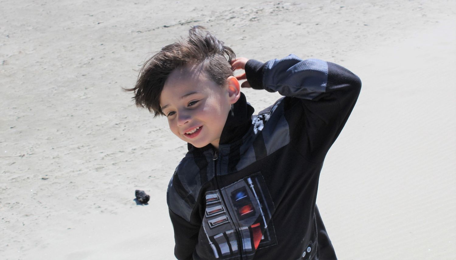 Boy wearing a Darth Vader costume jacket outdoors, smiling and posing with hand behind his head.