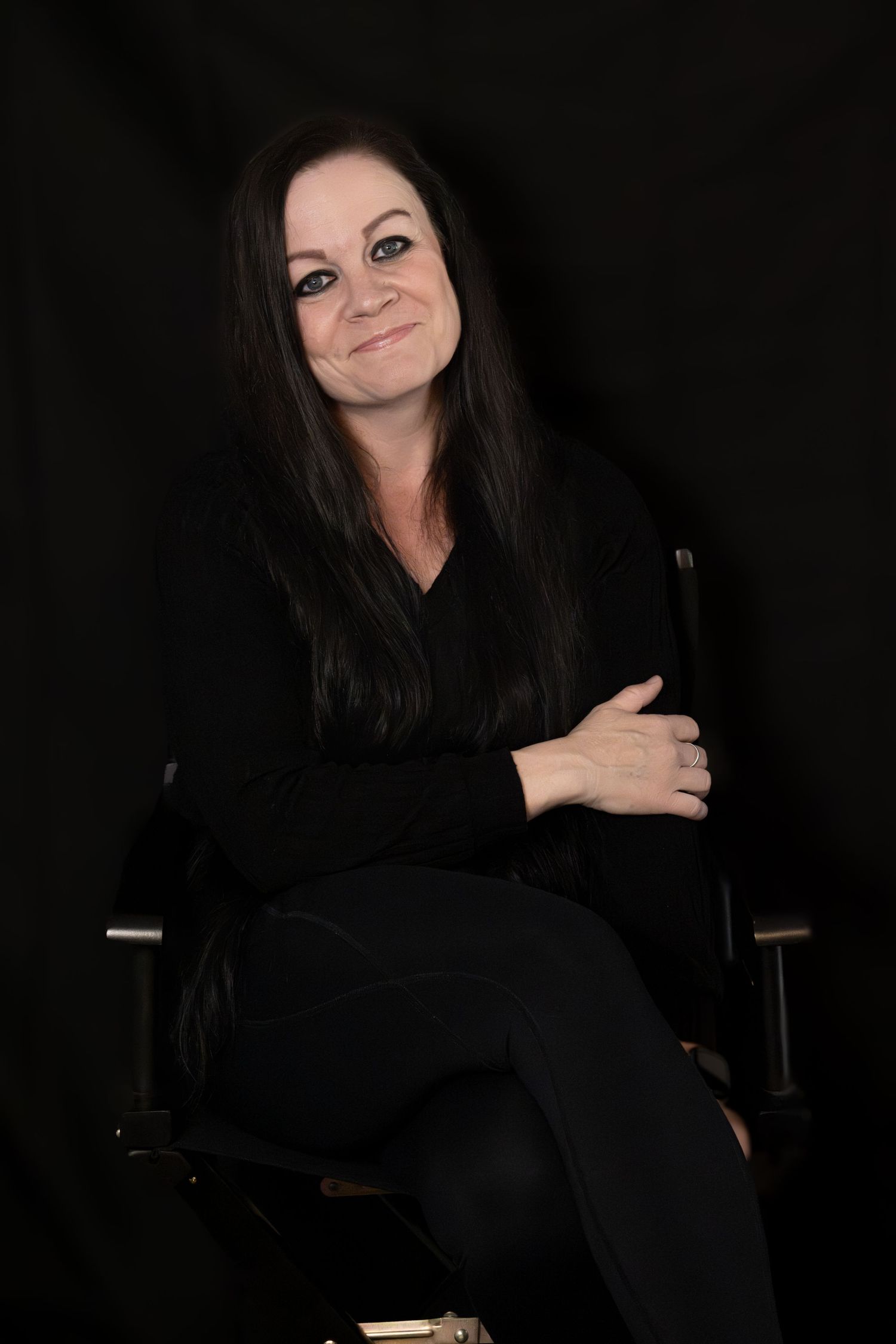 Woman in black outfit sitting in director's chair, smiling with arms crossed against dark background.