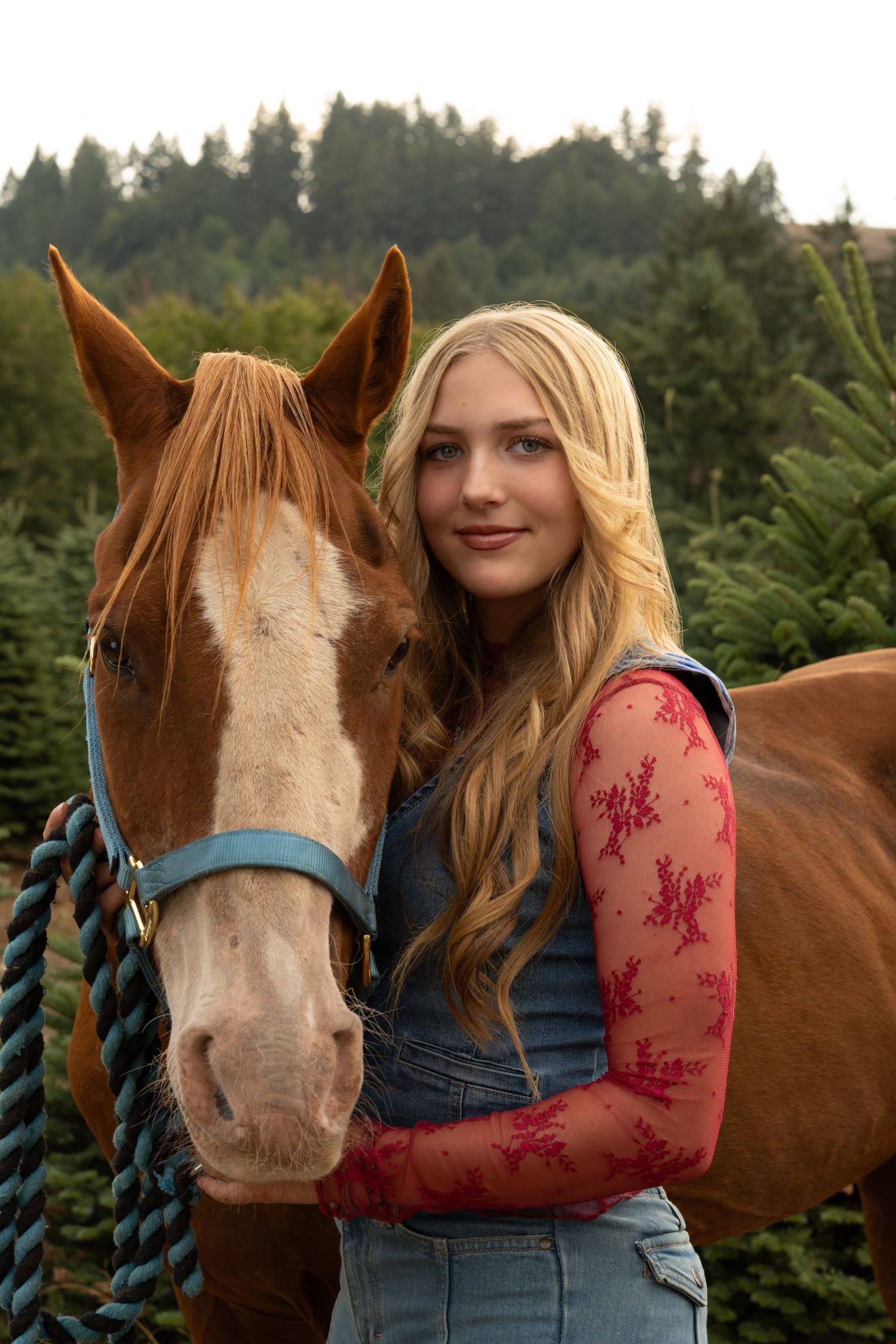 Young blonde woman in denim overalls and floral lace top posing with a chestnut horse in a Christmas tree farm.