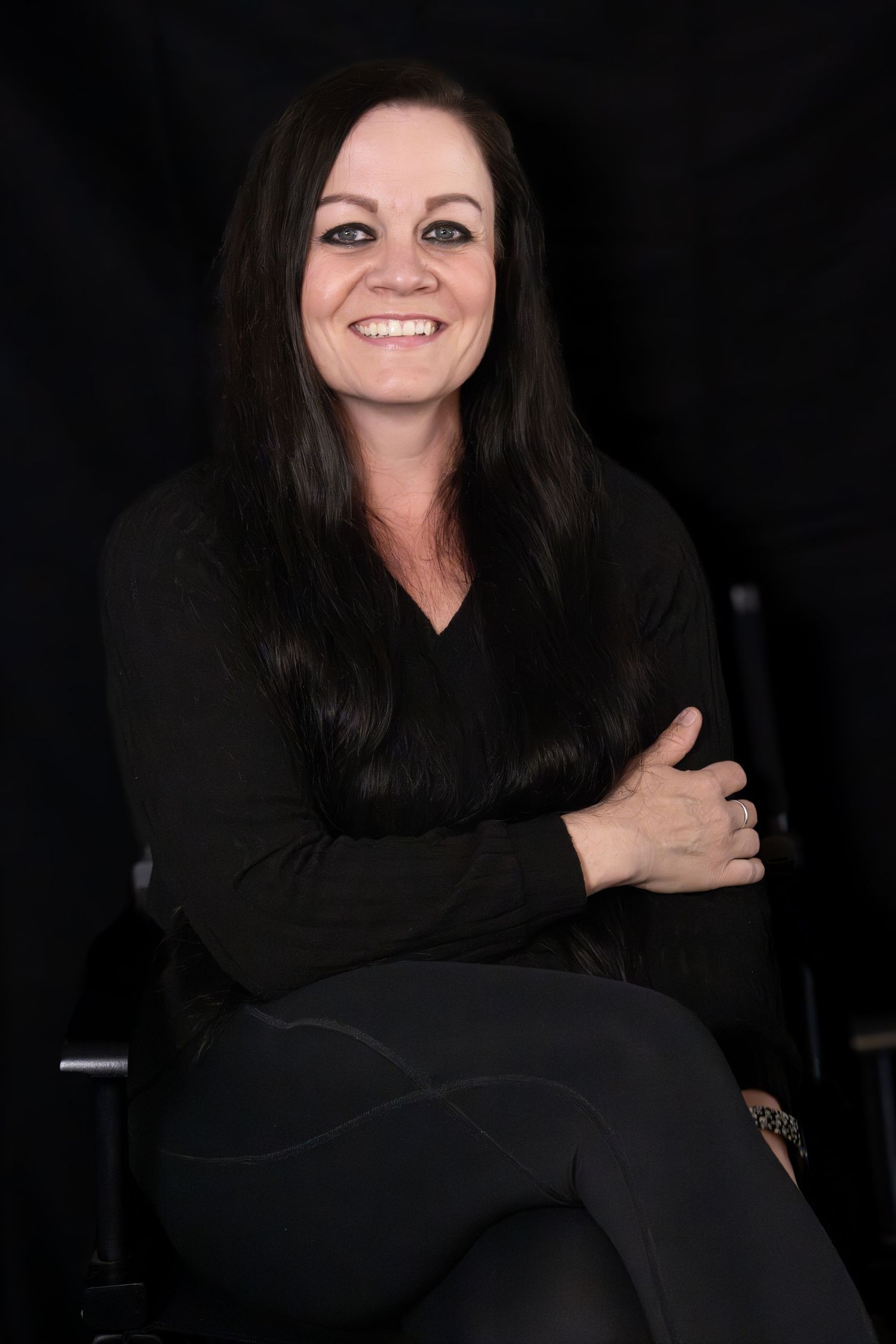 Smiling woman with dark hair in black outfit sits cross-armed against dark background.