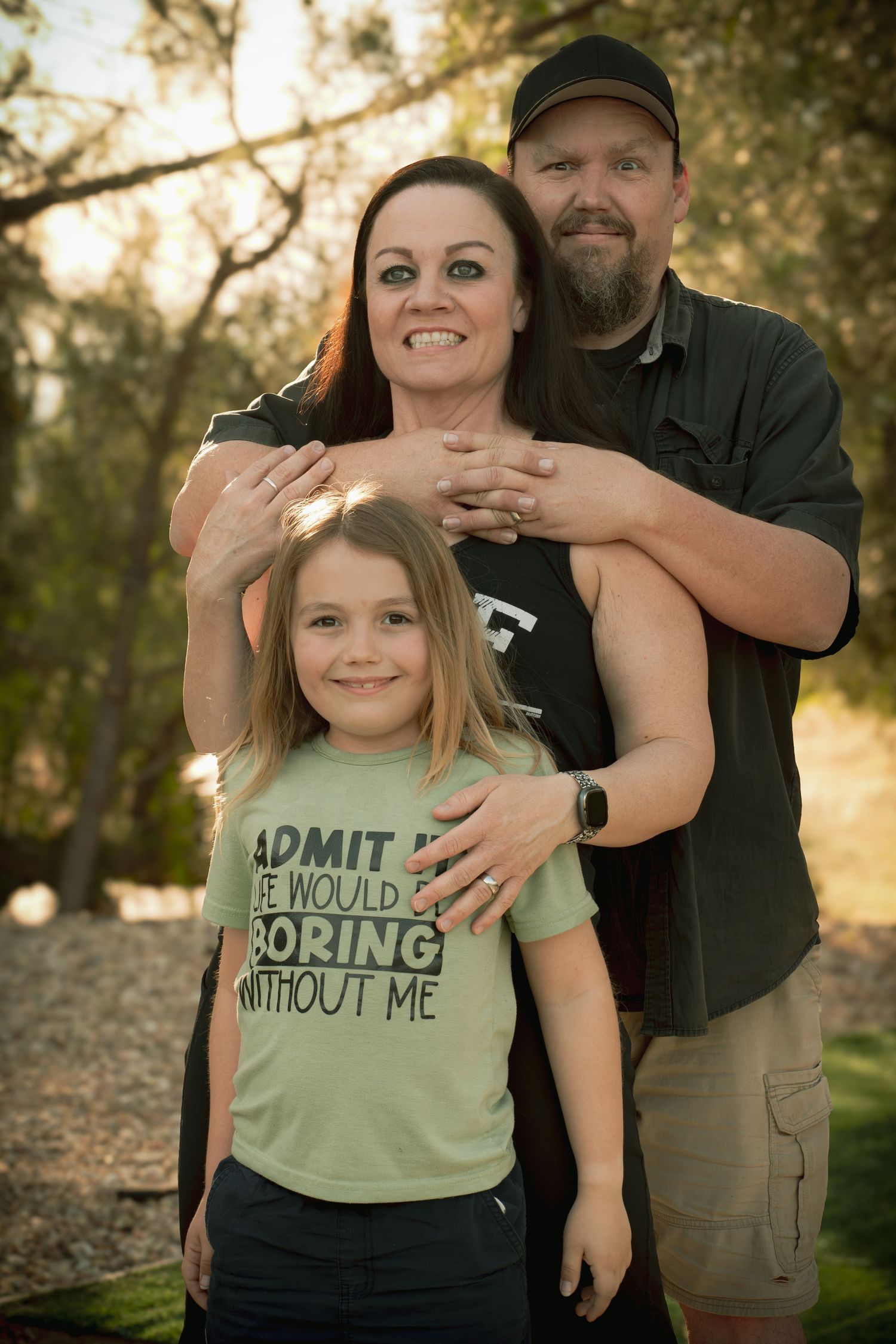 Family portrait of parents and young child in green shirt reading 'Admit it Life Would Be Boring Without Me' outdoors. making silly faces