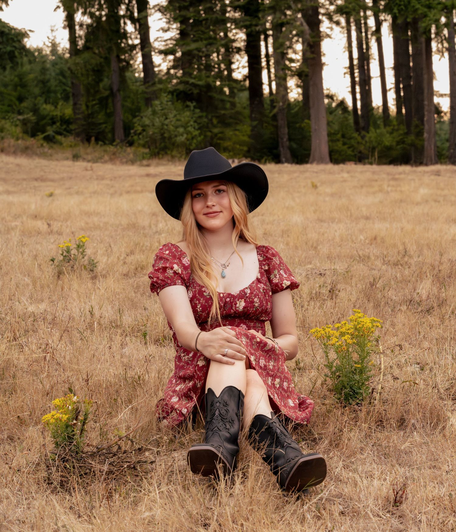 Blonde woman in floral dress, black cowboy hat and boots sits in golden meadow with tall pines.