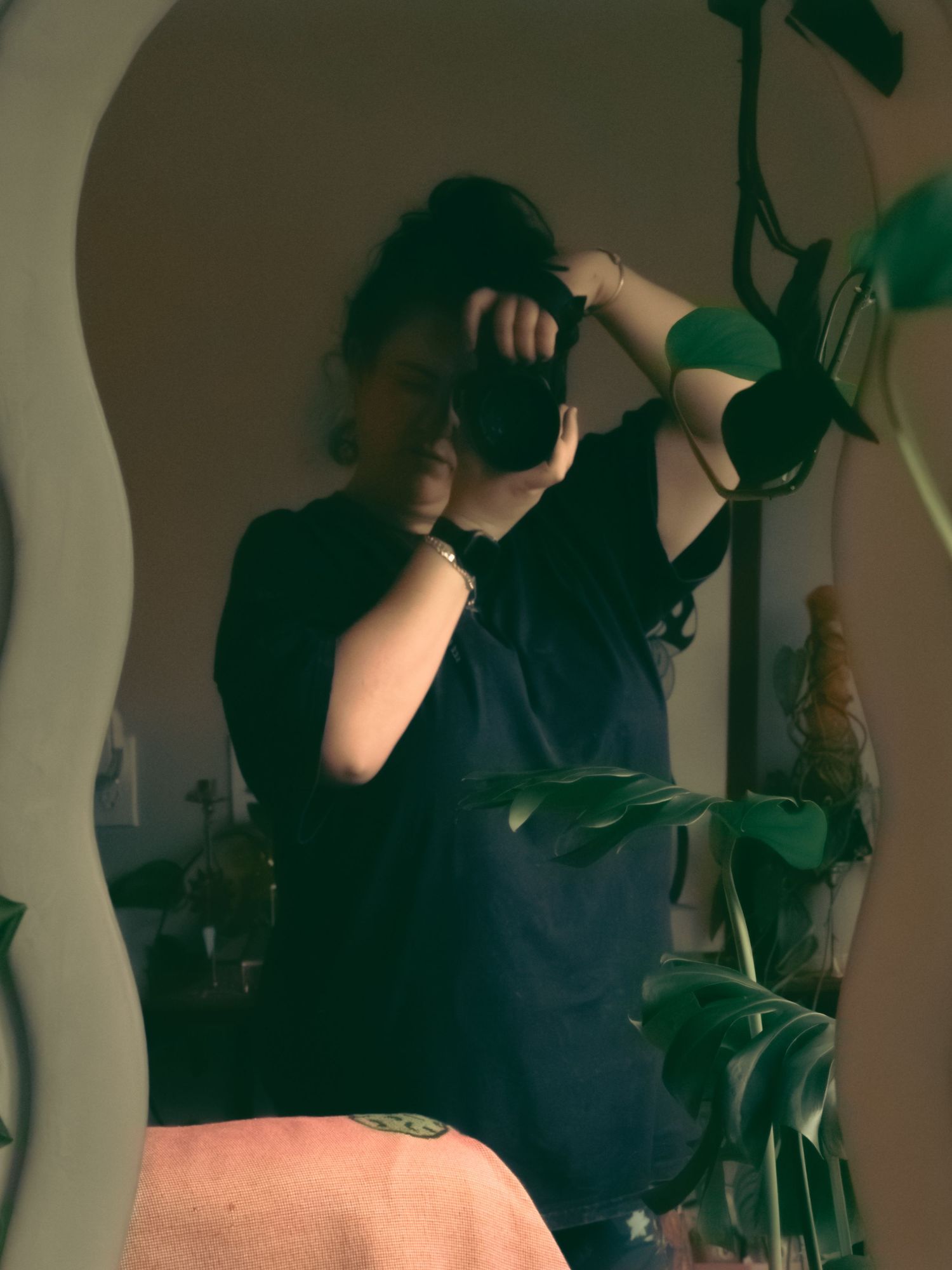 Ty Tofel a woman taking a mirror selfie with a  camera surrounded by indoor houseplants in dim light. earthy,moody vibe