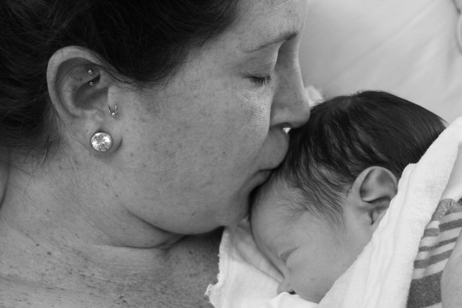 Black and white photo of a mother tenderly kissing her newborn baby wrapped in a white blanket.