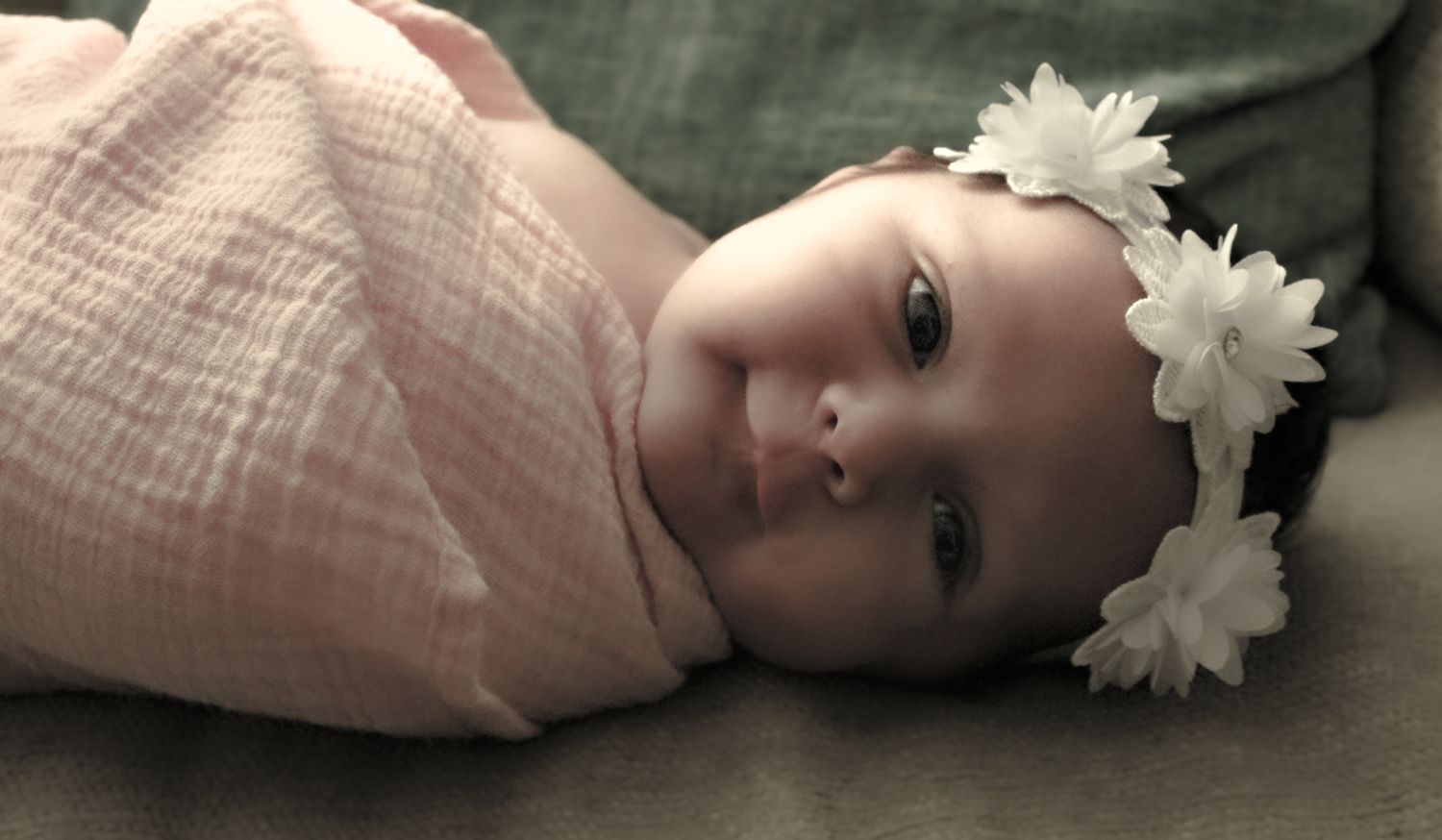Smiling baby girl wrapped in pink swaddle blanket wearing white flower headband lying on soft surface.