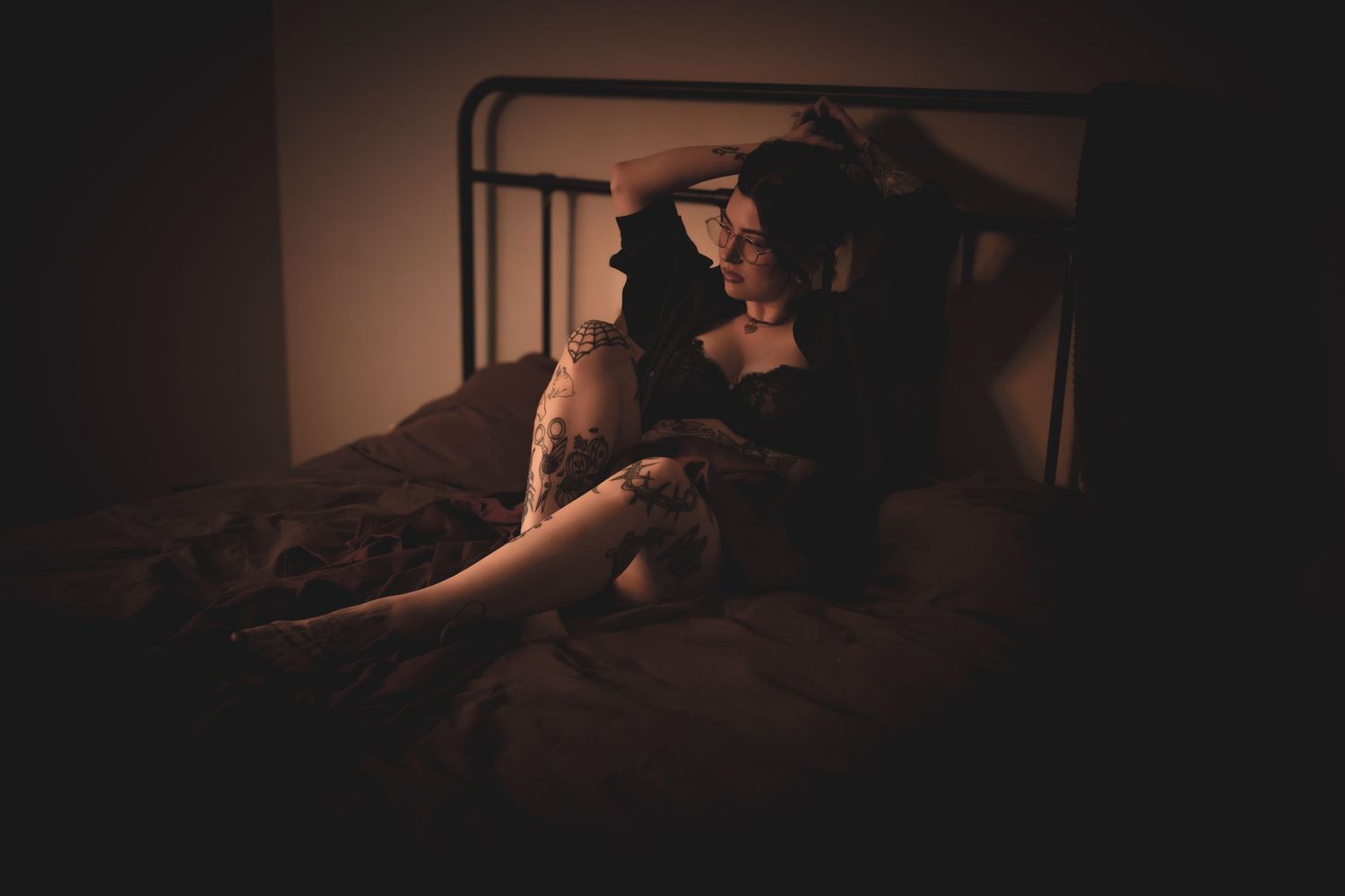 Woman with tattoos posing on bed in dim warm lighting in lingerie.
