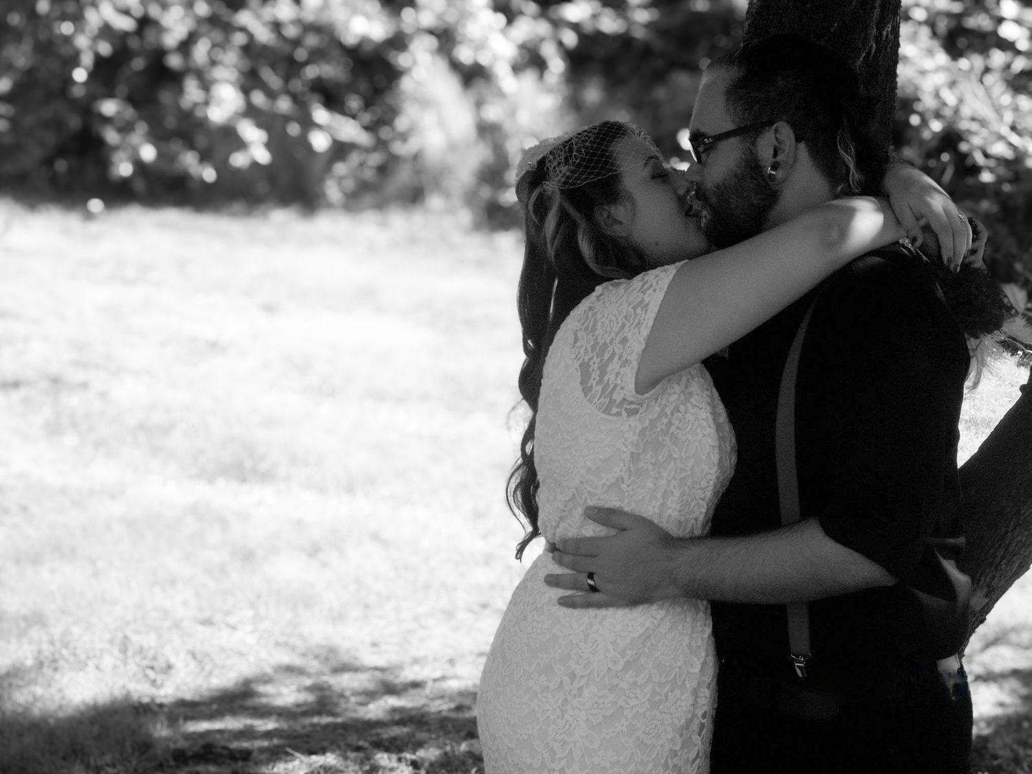 Black and white photo of a couple sharing a romantic kiss outdoors, bride in lace dress embracing groom near a tree.