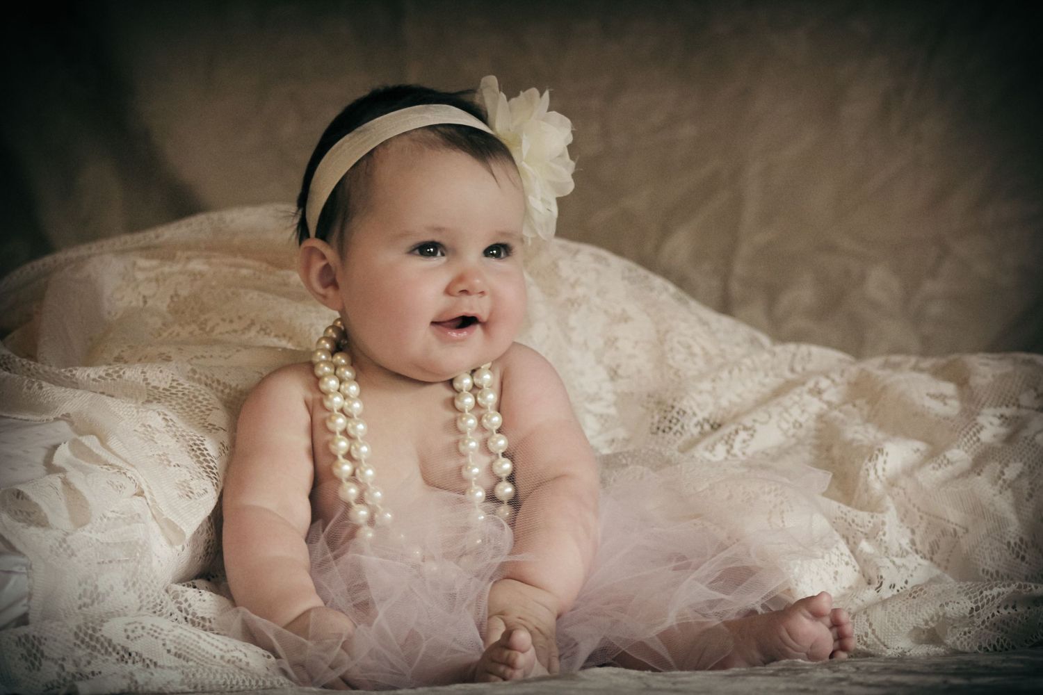 Adorable baby girl wearing pearl necklaces and a floral headband, sitting on lace fabric in a soft vintage-style portrait.