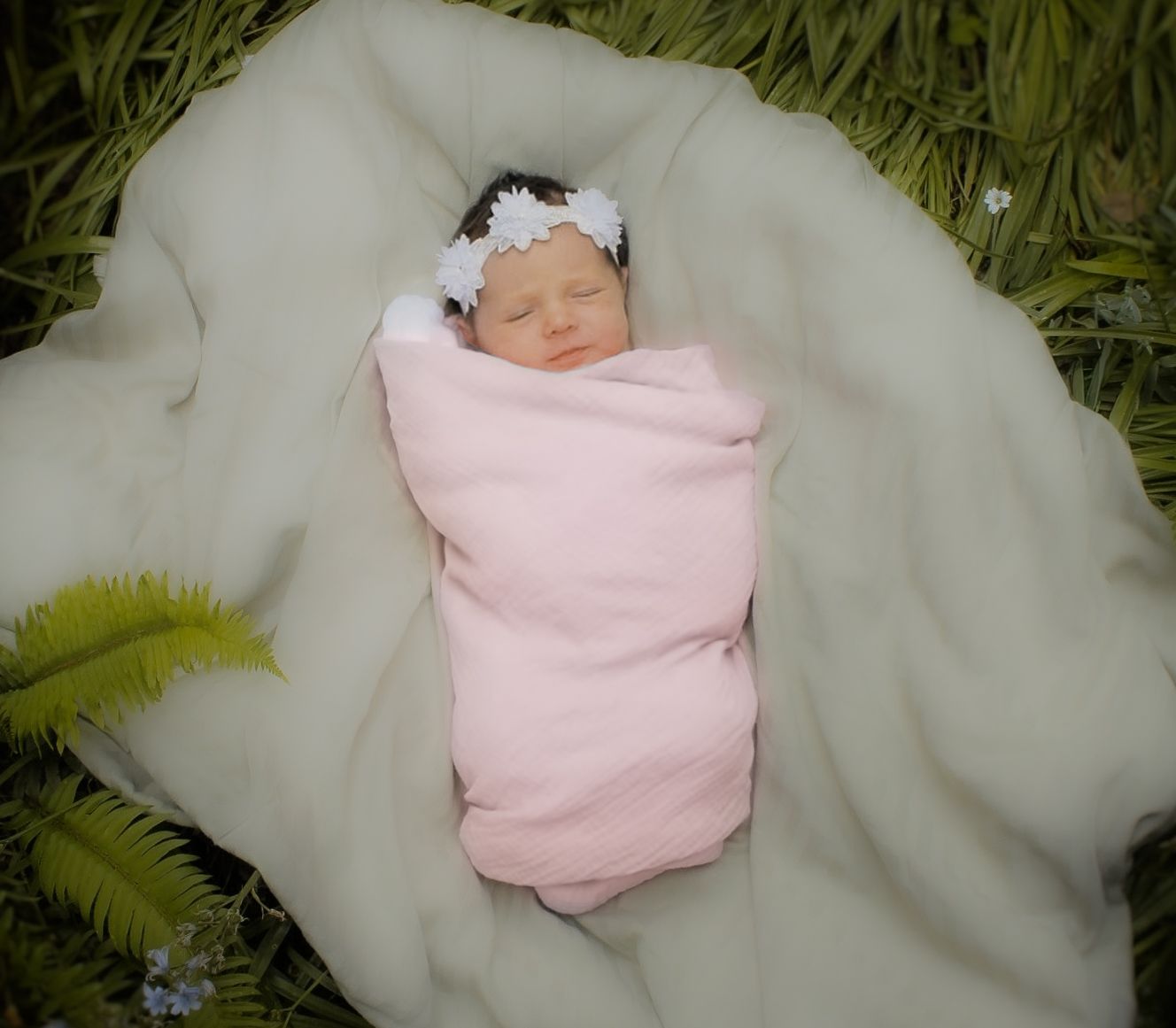 Sleeping newborn baby girl swaddled in pink blanket with white daisy flower headband outdoors.