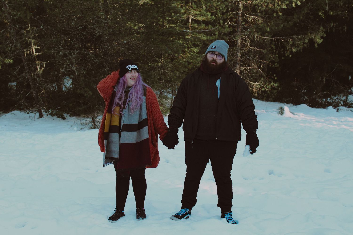 Couple holding hands in snowy winter landscape, wearing colorful cold weather gear with evergreen trees behind them.