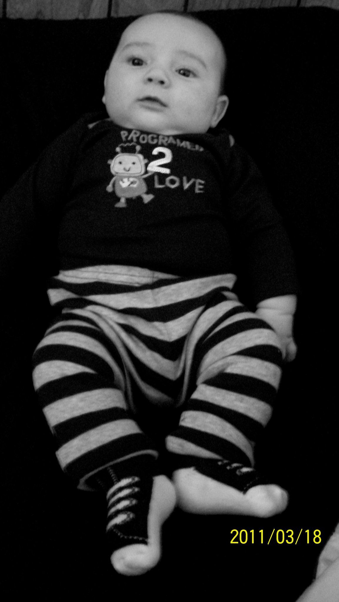 Black and white photo of a baby wearing striped pants and a Programmed 2 Love shirt, lying down, dated 2011/03/18.