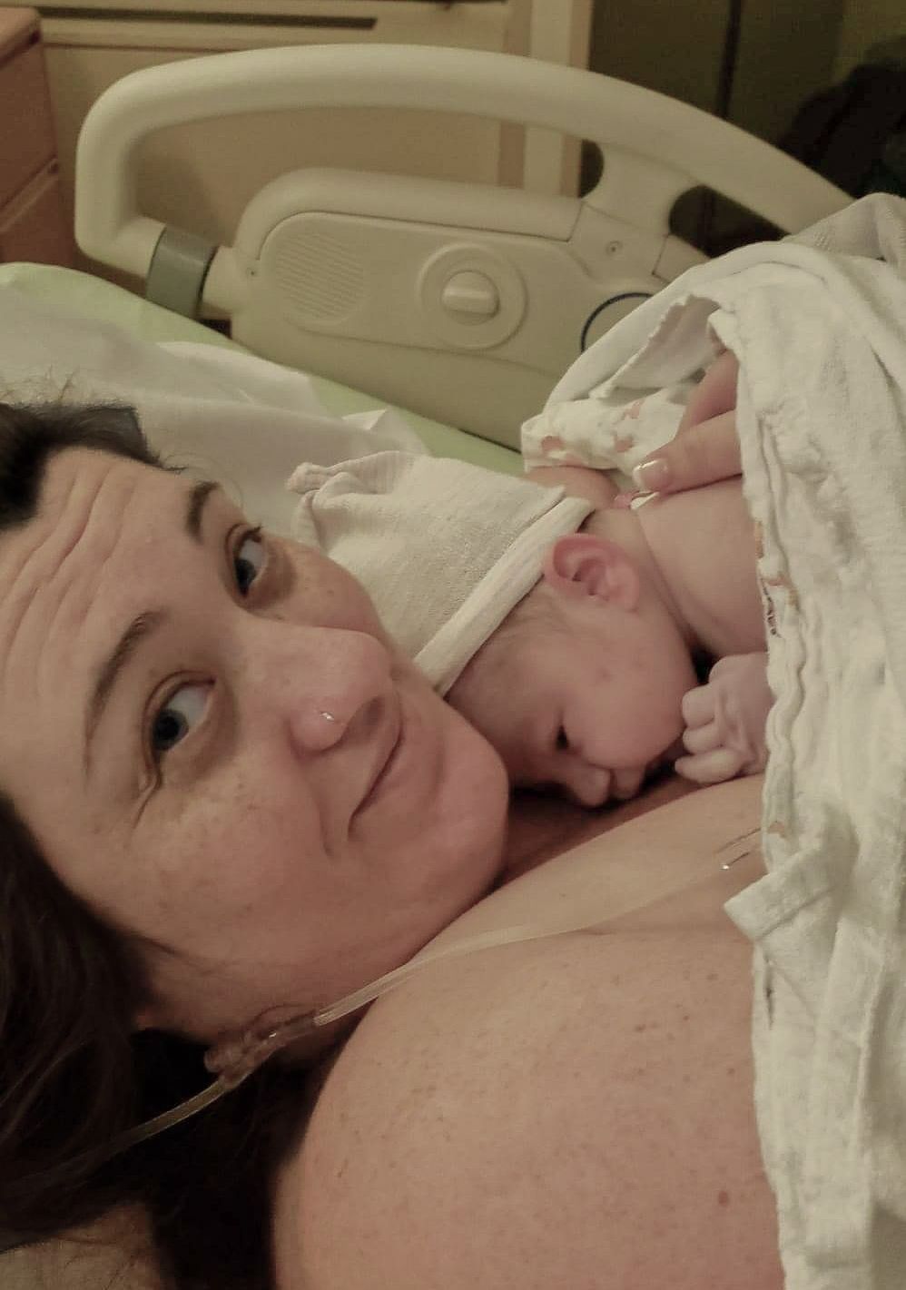 New mother lying in hospital bed smiling while holding her newborn baby wrapped in white blanket.