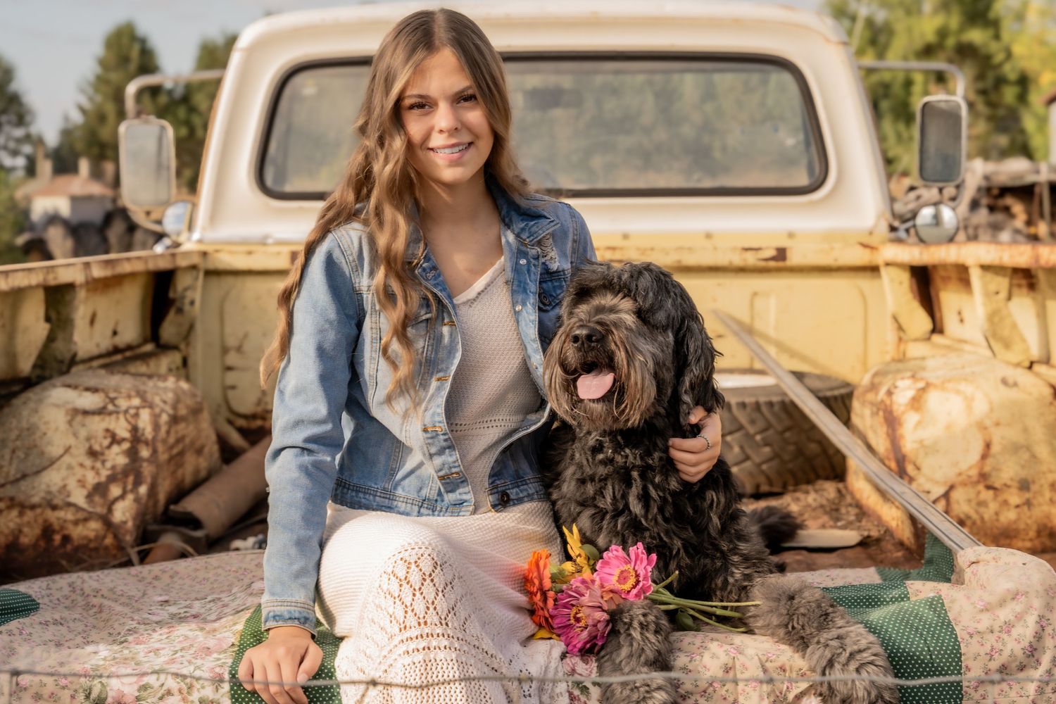 Young woman in denim jacket smiles beside a black dog in a vintage truck bed, holding colorful flowers.