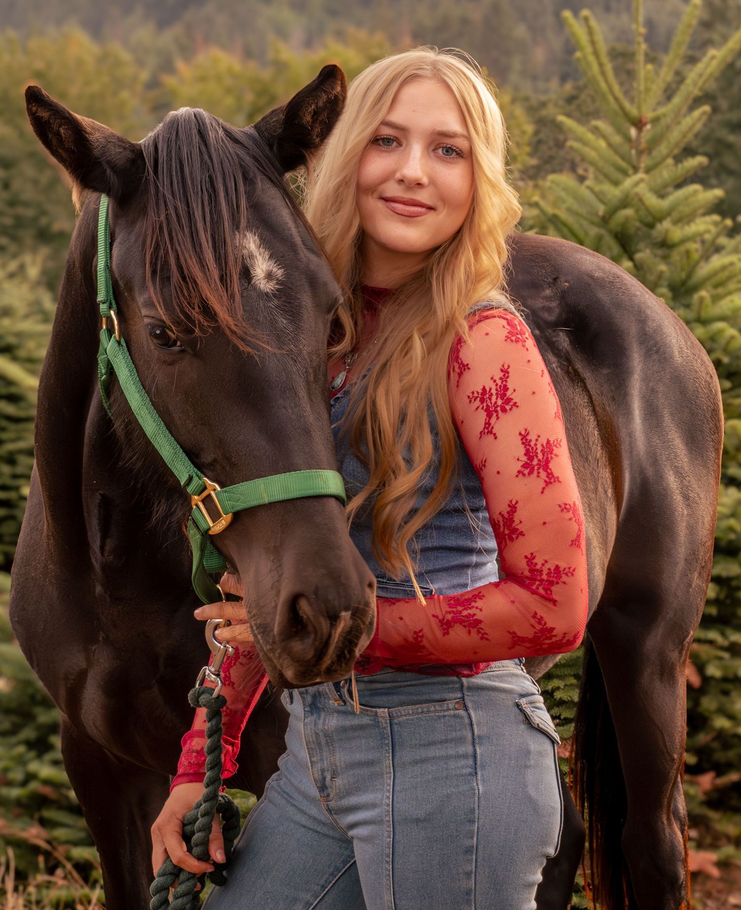 Young blonde woman in floral top and jeans poses with a dark horse wearing green halter among evergreen trees.