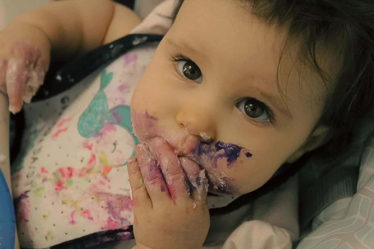 Messy baby eating colorful food with purple-stained hands and bib, sitting in a high chair.