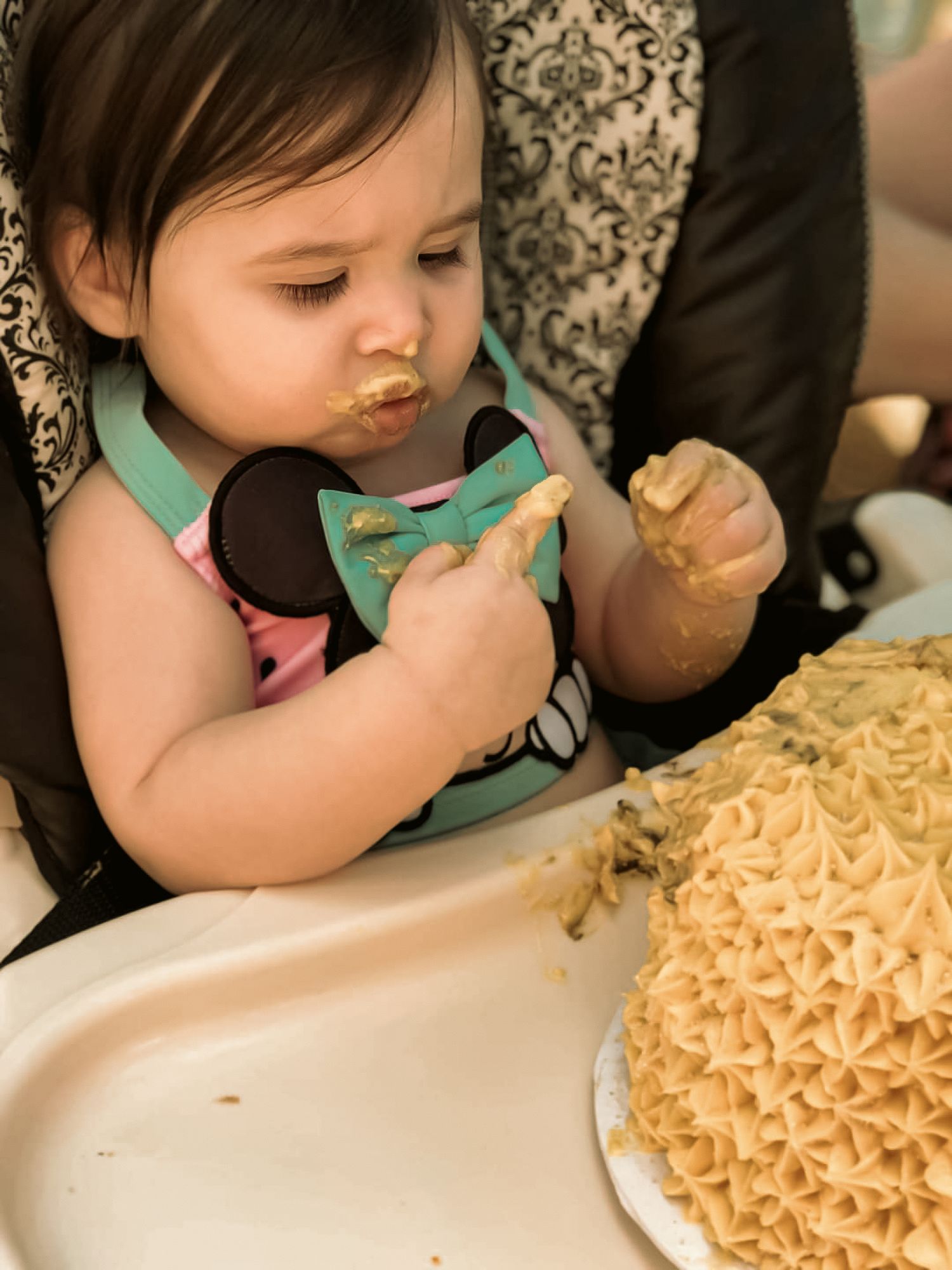 Baby in high chair eagerly eating smash cake with frosting on face during first birthday celebration.