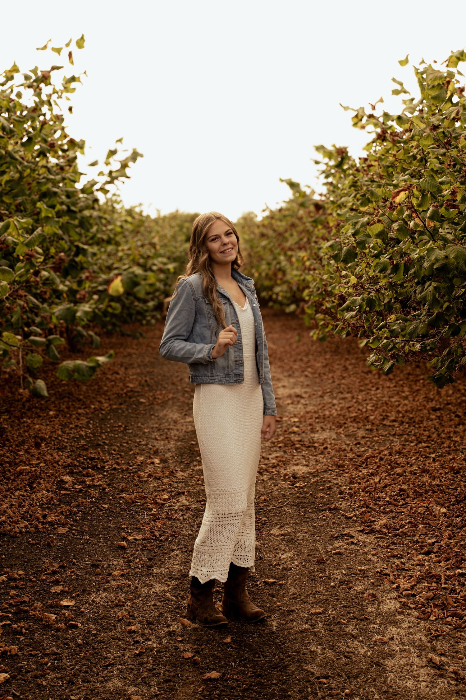 Young woman in white lace dress and denim jacket posing in an orchard pathway with lush green trees.