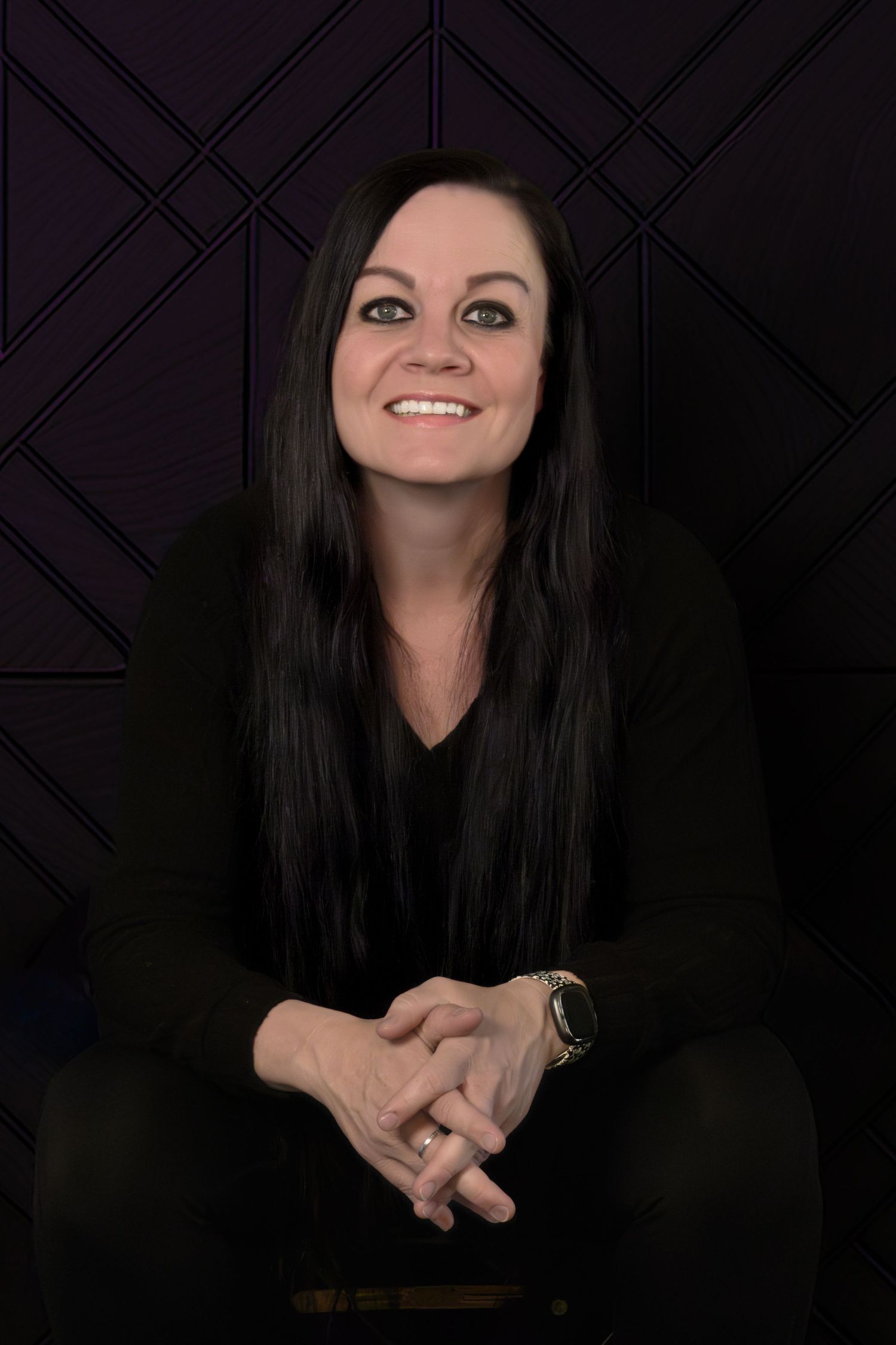 Smiling woman with long dark hair wearing black outfit, seated with hands clasped against dark geometric patterned background.