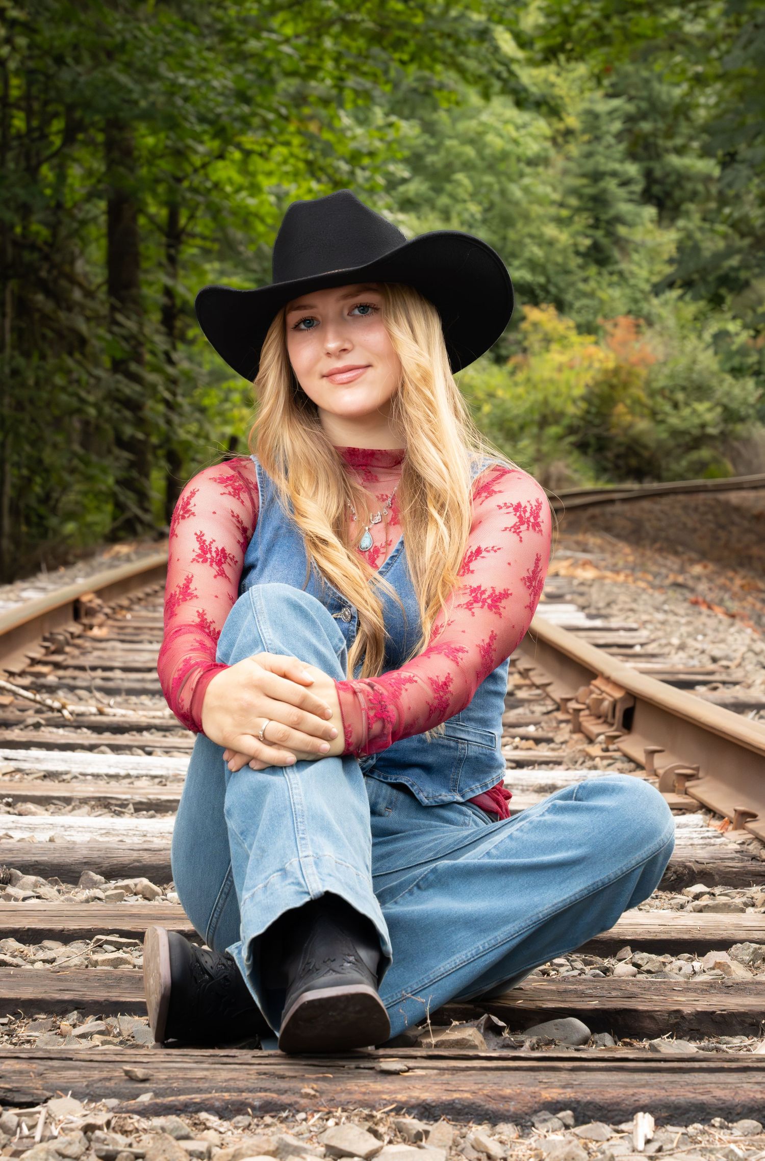 Young blonde woman in black cowboy hat, denim overalls, and red lace top sitting on railroad tracks in a forest. lyons OR