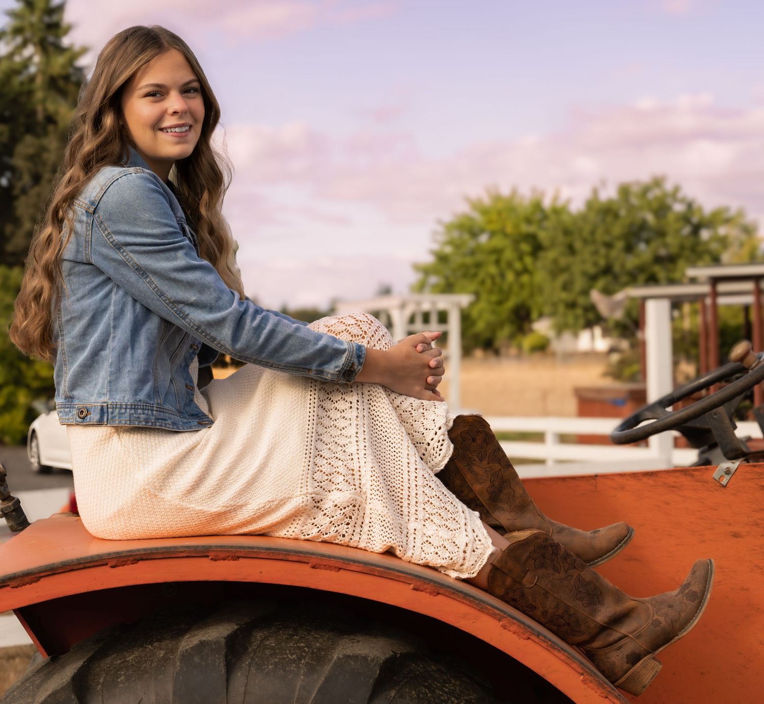 Young woman in denim jacket, white knit dress and cowboy boots sitting on a tractor in a farm setting at sunset. crabtree oregon