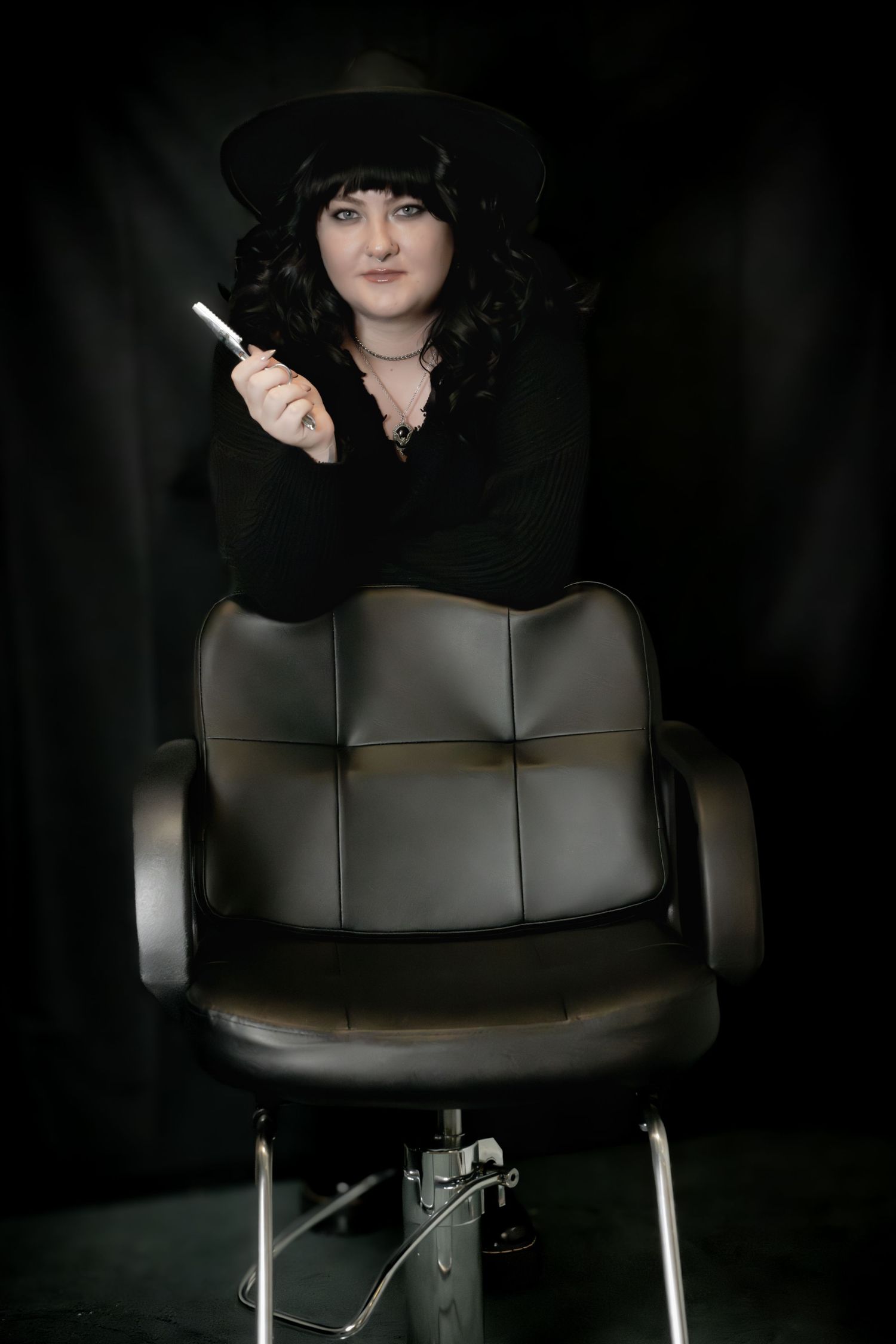 Woman in black hat and outfit holding cigarette, leaning on leather salon chair against dark background.