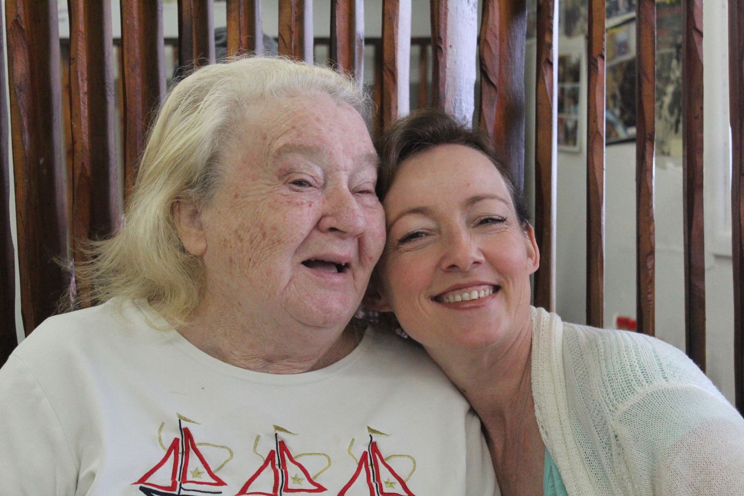 Two women smiling and posing cheek-to-cheek, one elderly in a sailboat shirt, one middle-aged, indoors.