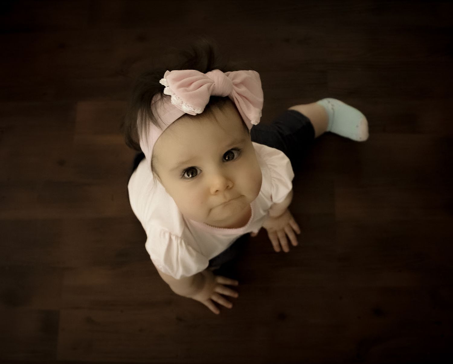 Adorable baby girl in pink bow headband and white outfit looks up at camera on dark wood floor.