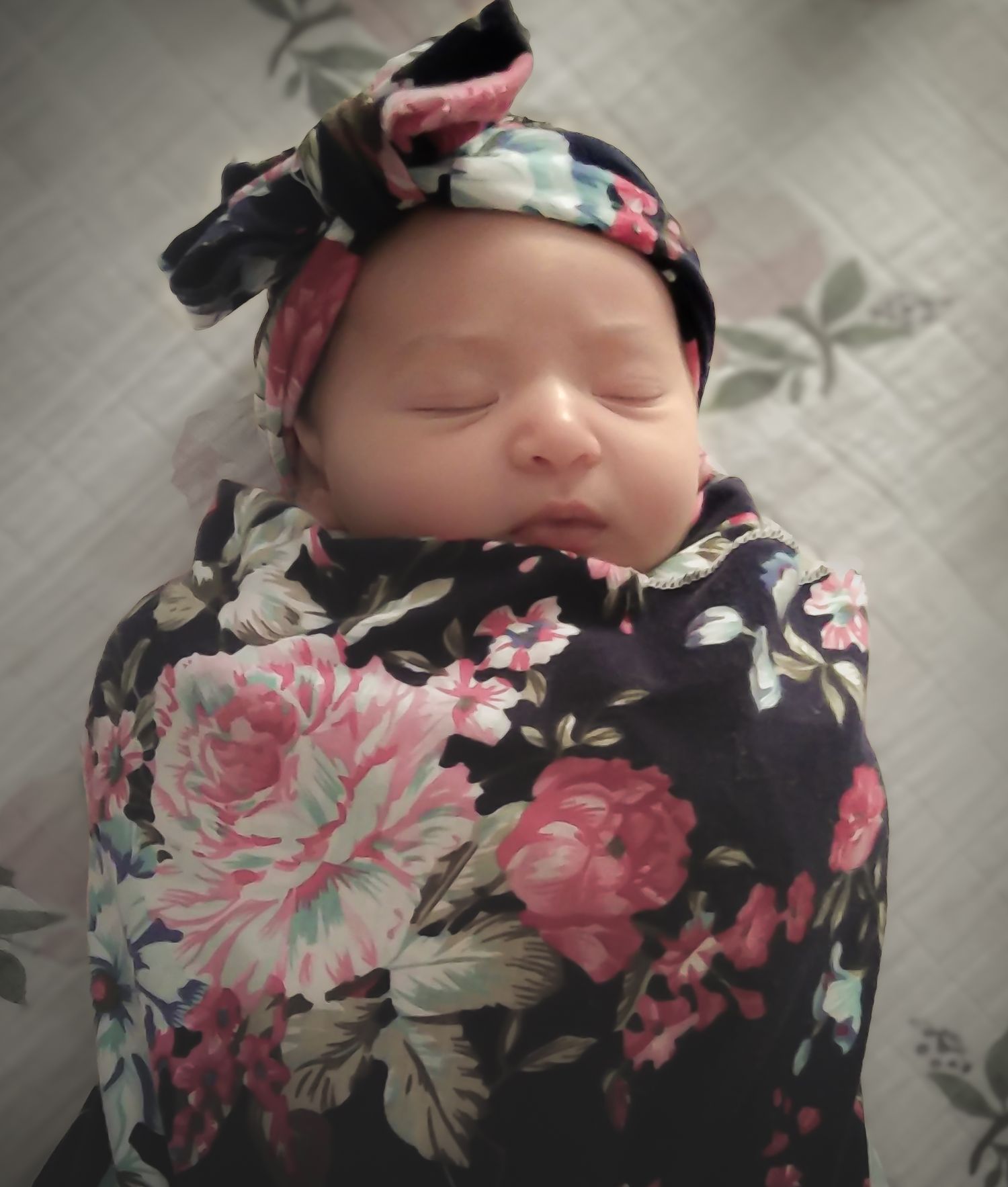 Newborn baby sleeping peacefully swaddled in a dark floral blanket with matching bow headband.