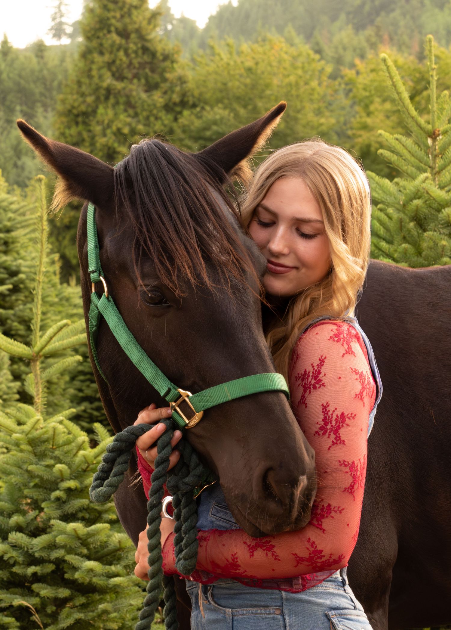 Blonde girl hugging a dark brown horse wearing a jean vest over a red lace long sleeve shirt, surrounded by lush green pine trees. lyons OR
