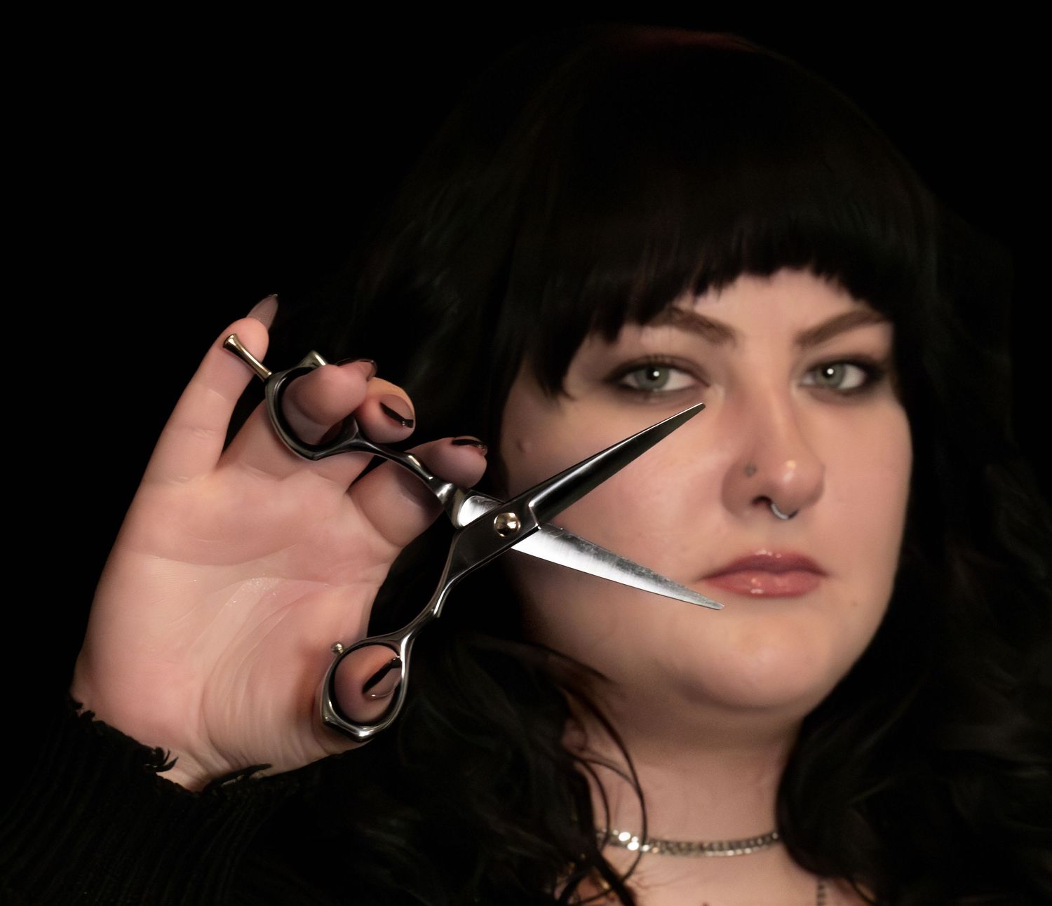Woman with black hair holding scissors close to camera, styled as a professional hairstylist against dark background business branding