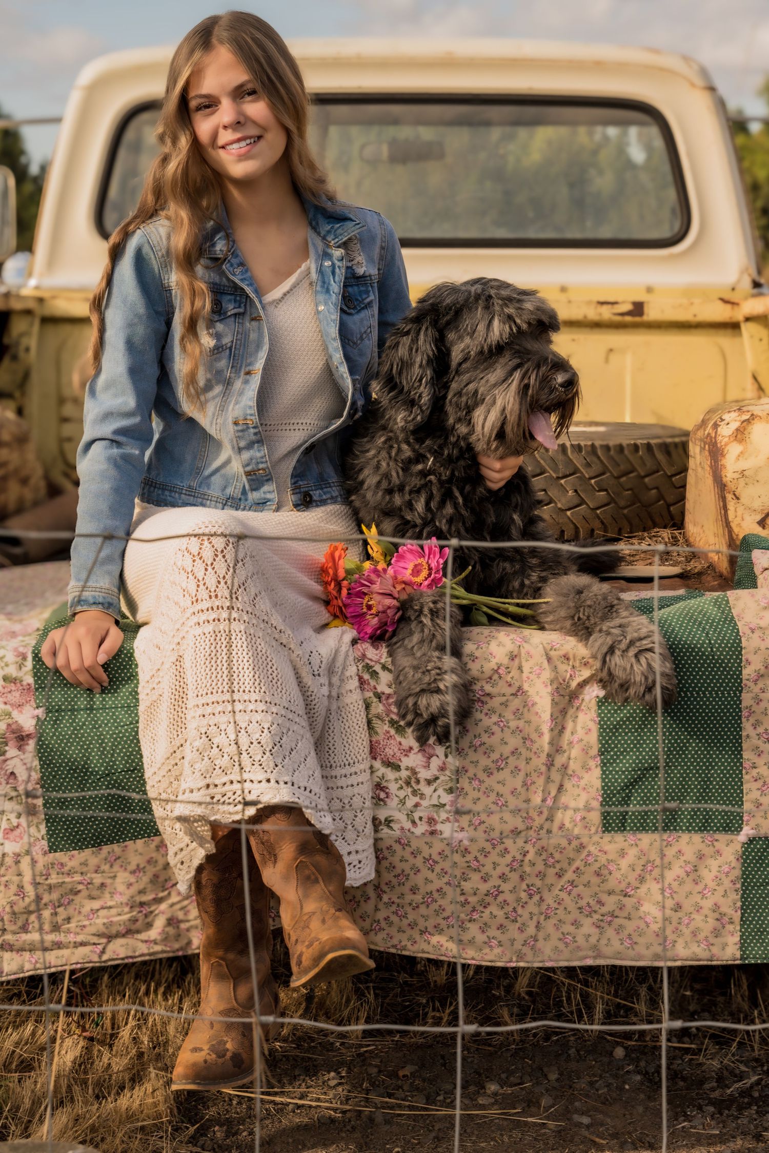 Young woman in denim jacket and cowboy boots sits on vintage truck tailgate with black fluffy dog and flowers.