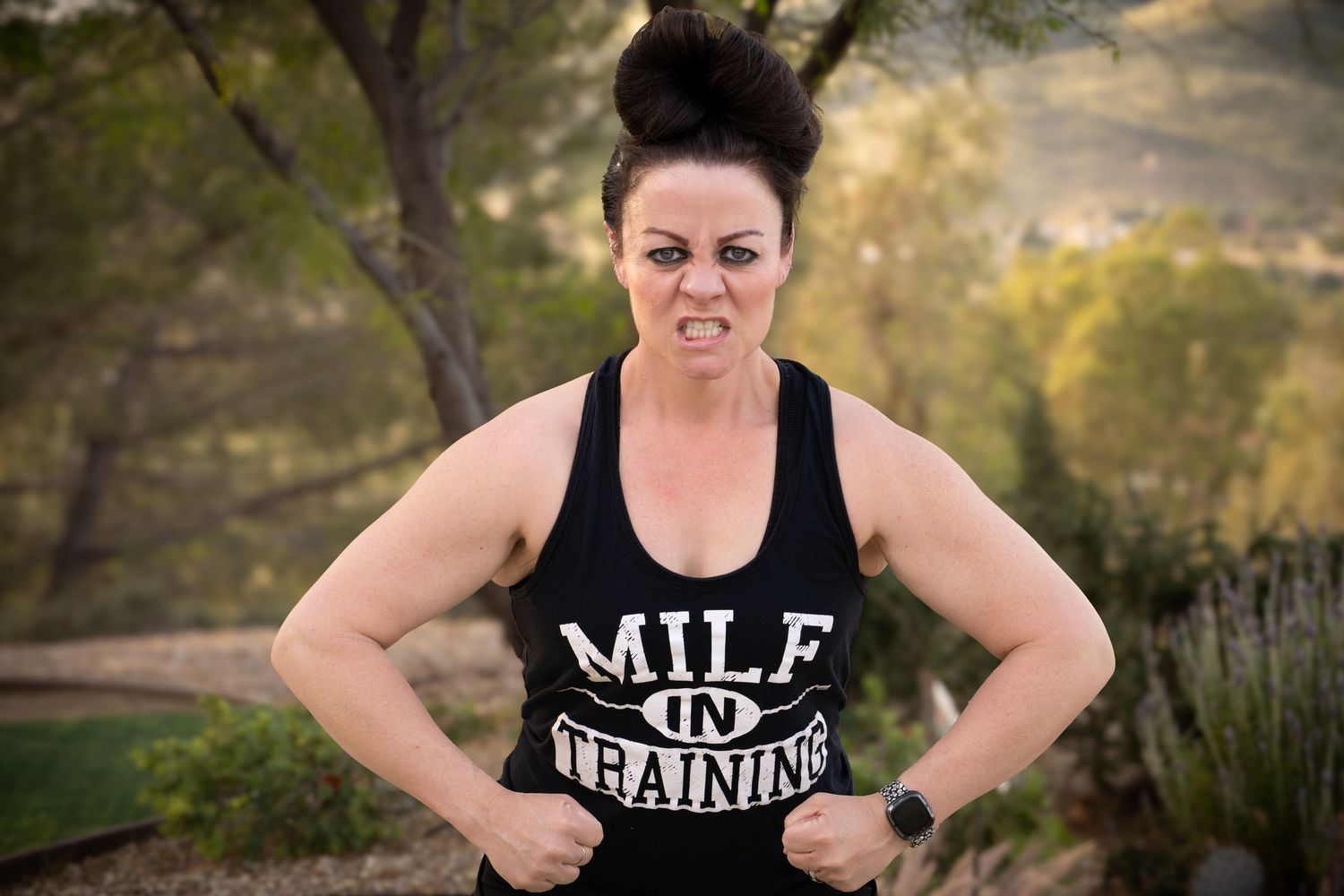 Woman in black MILF in Training tank top flexing outdoors with trees in background.