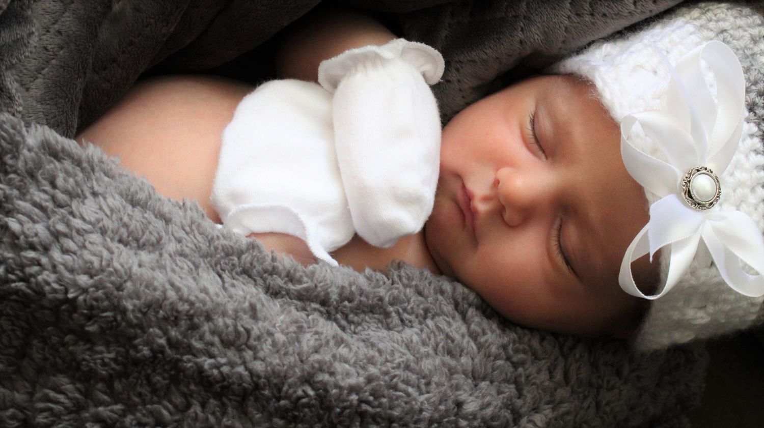 Peaceful newborn baby sleeping on a soft gray blanket wearing a white outfit and white bow headband.