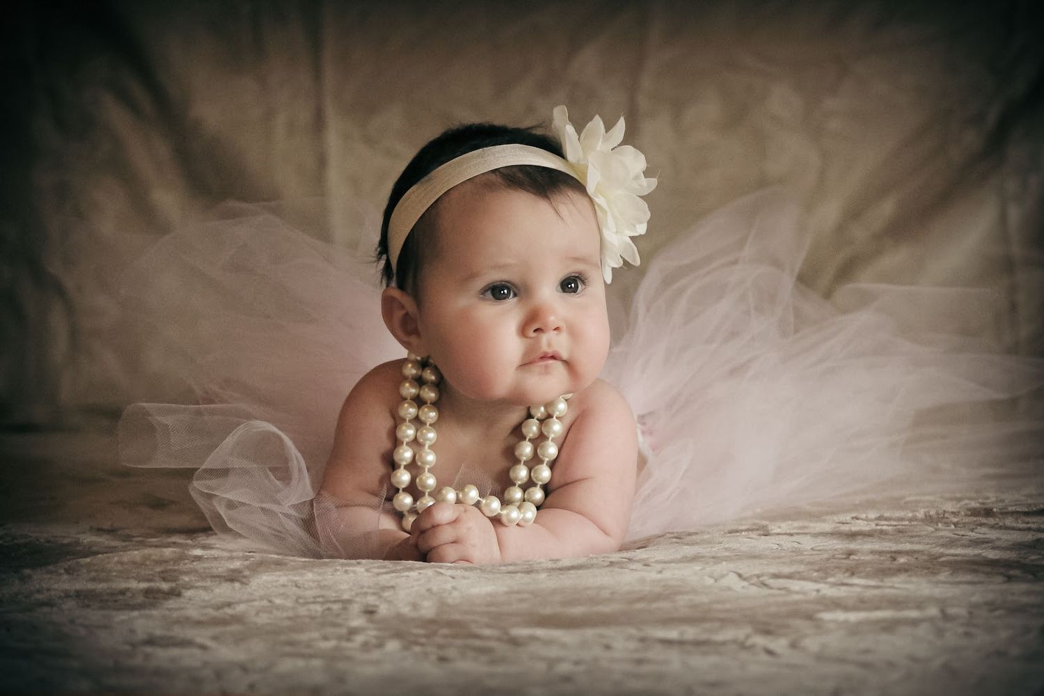 Adorable baby girl wearing pearl necklace, floral headband, and tutu, posing on a vintage lace backdrop.