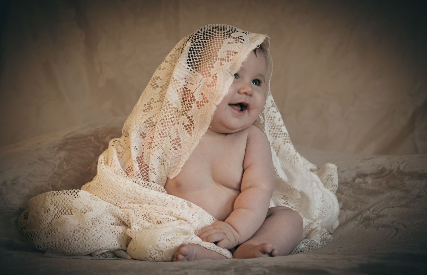 Adorable baby wrapped in delicate lace fabric smiling, captured in a soft vintage-style portrait photography setting.