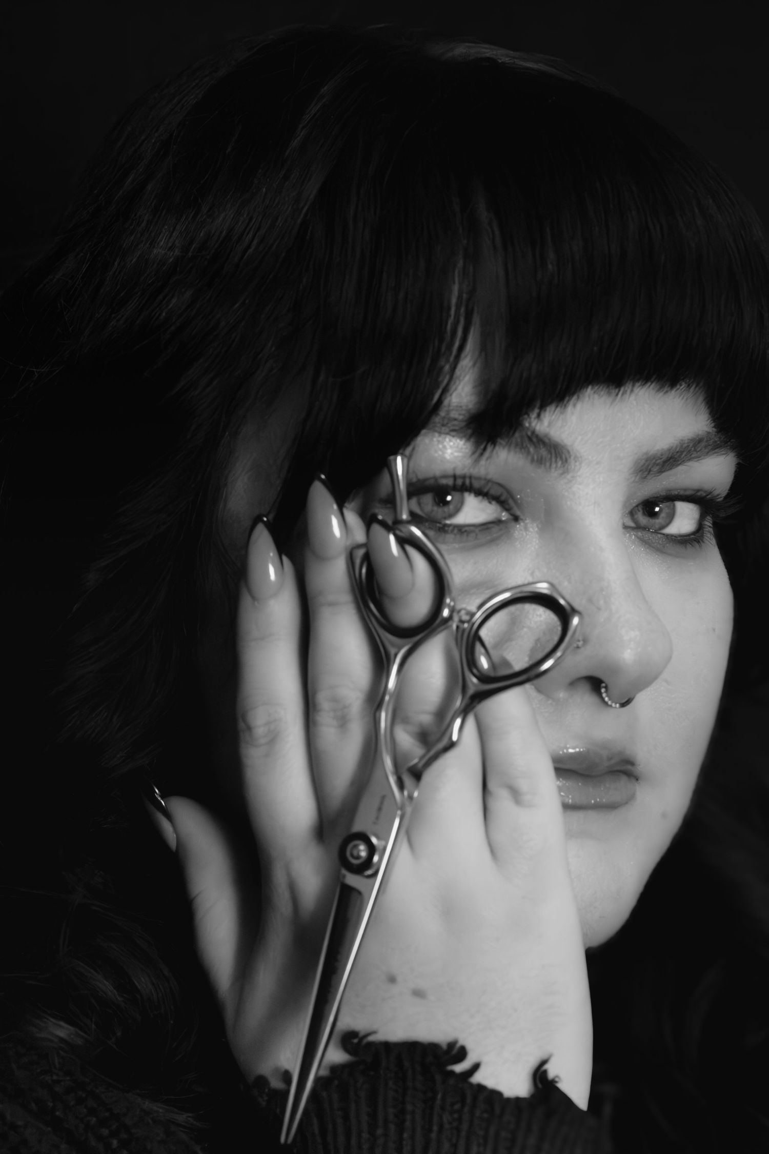 Black and white portrait of a woman with bangs holding hairdressing scissors near her face.