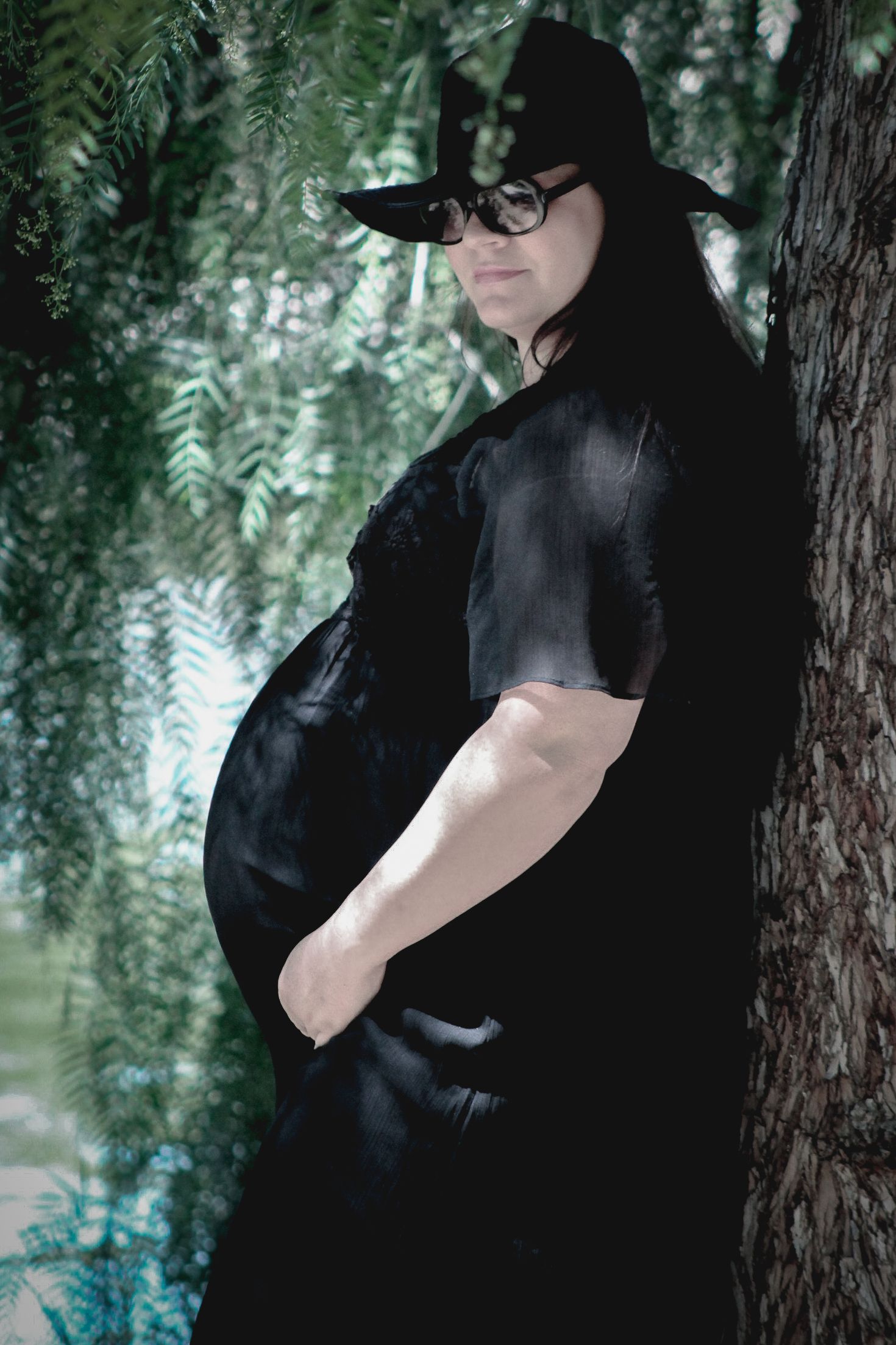 Pregnant woman in black witch costume and hat leaning against tree outdoors.