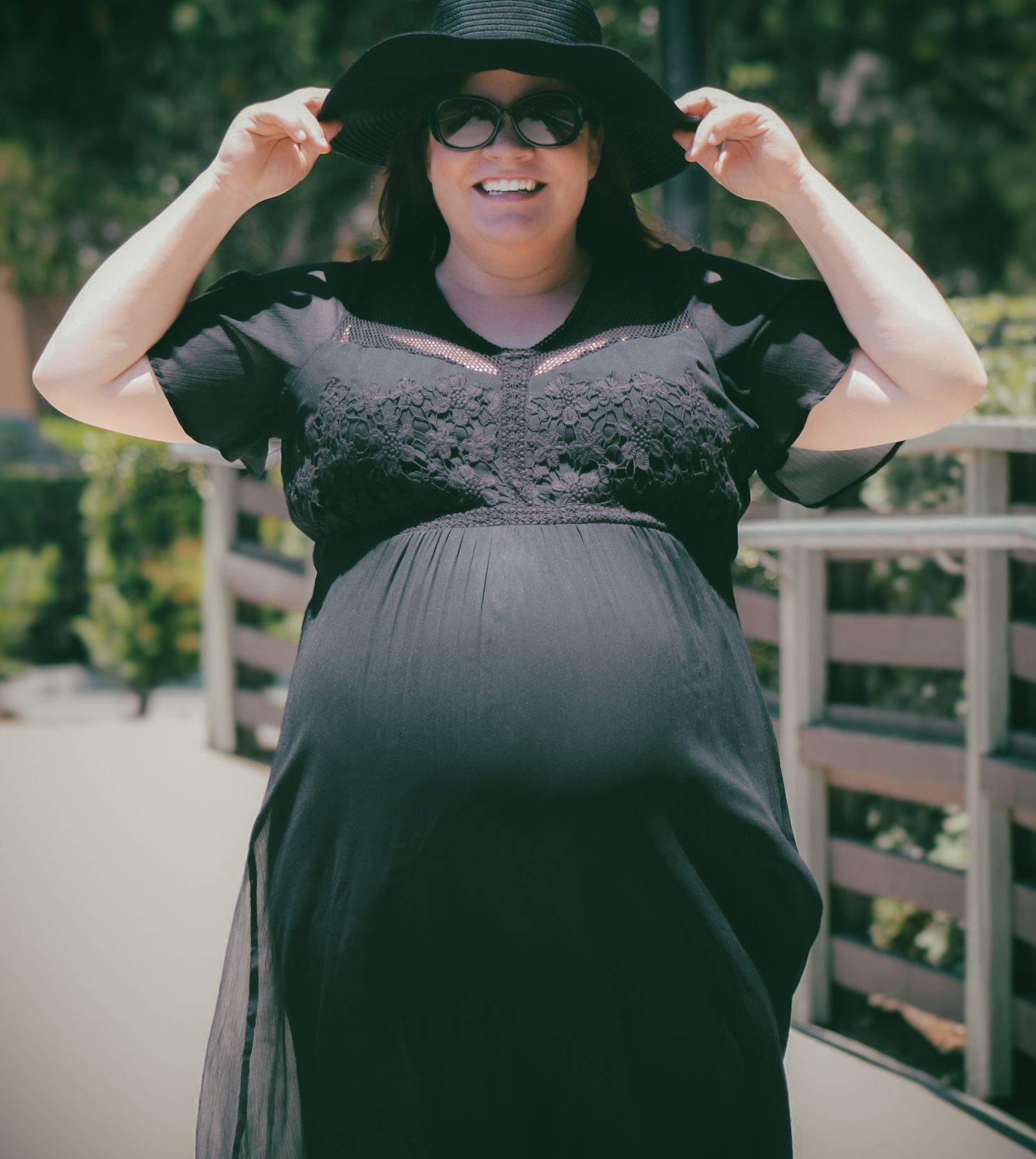 Pregnant woman in black dress and wide-brim hat smiling outdoors on sunny day.