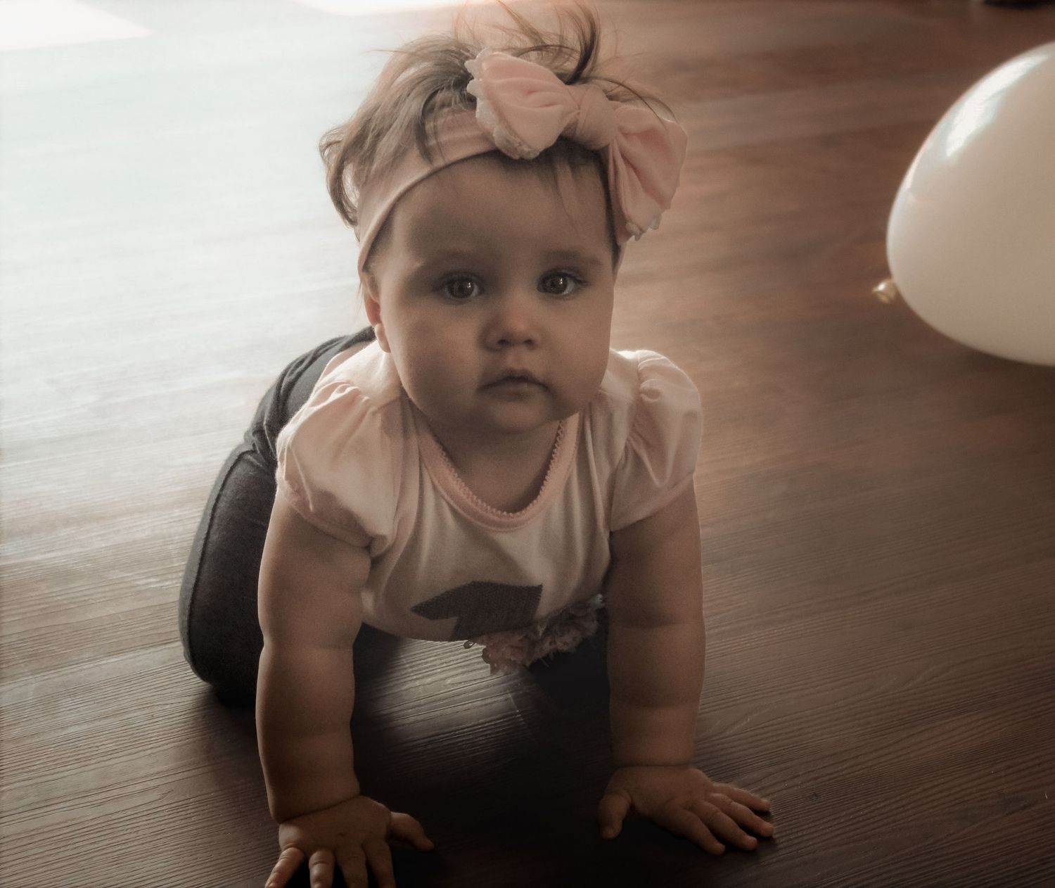 Adorable baby girl in pink bow headband crawling on hardwood floor with a white balloon in the background.