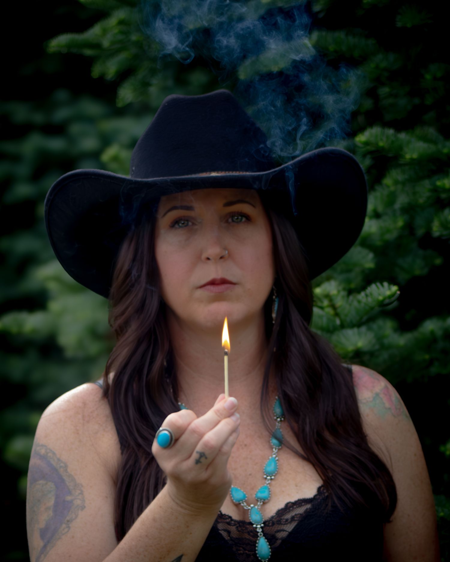 Woman in black cowboy hat holding lit match with smoke rising outdoors.