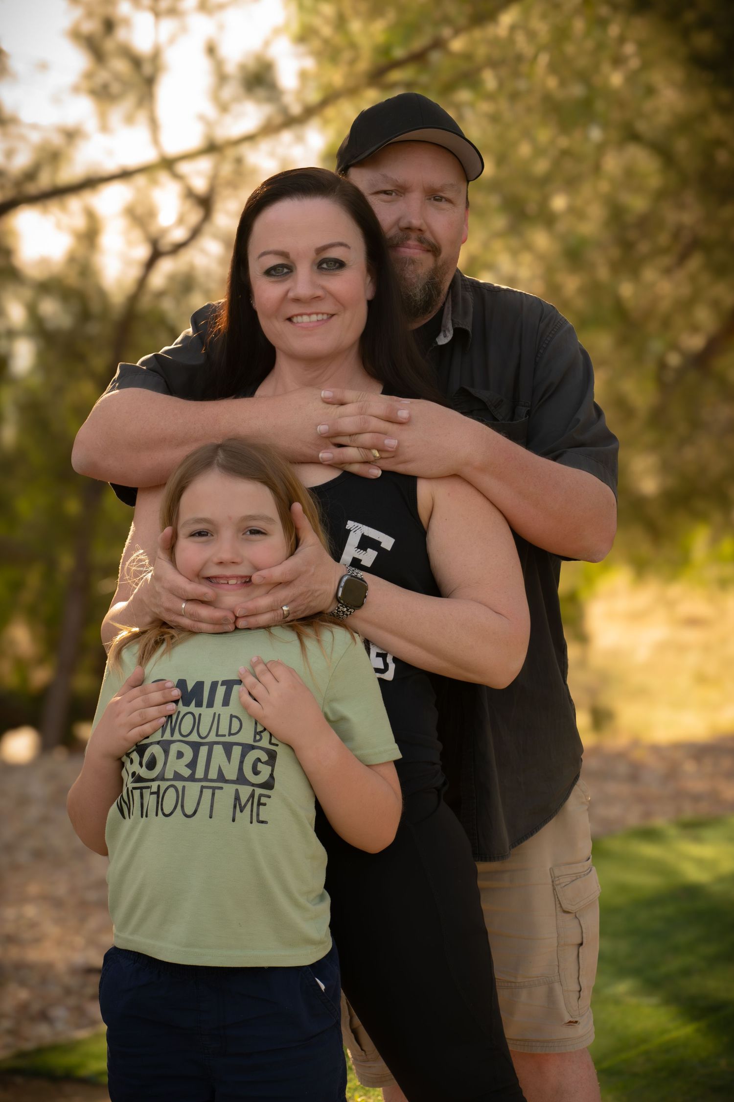 A smiling family of three poses outdoors, with a man hugging a woman and young boy in a golden hour sunset setting. Authentic moody outdoor