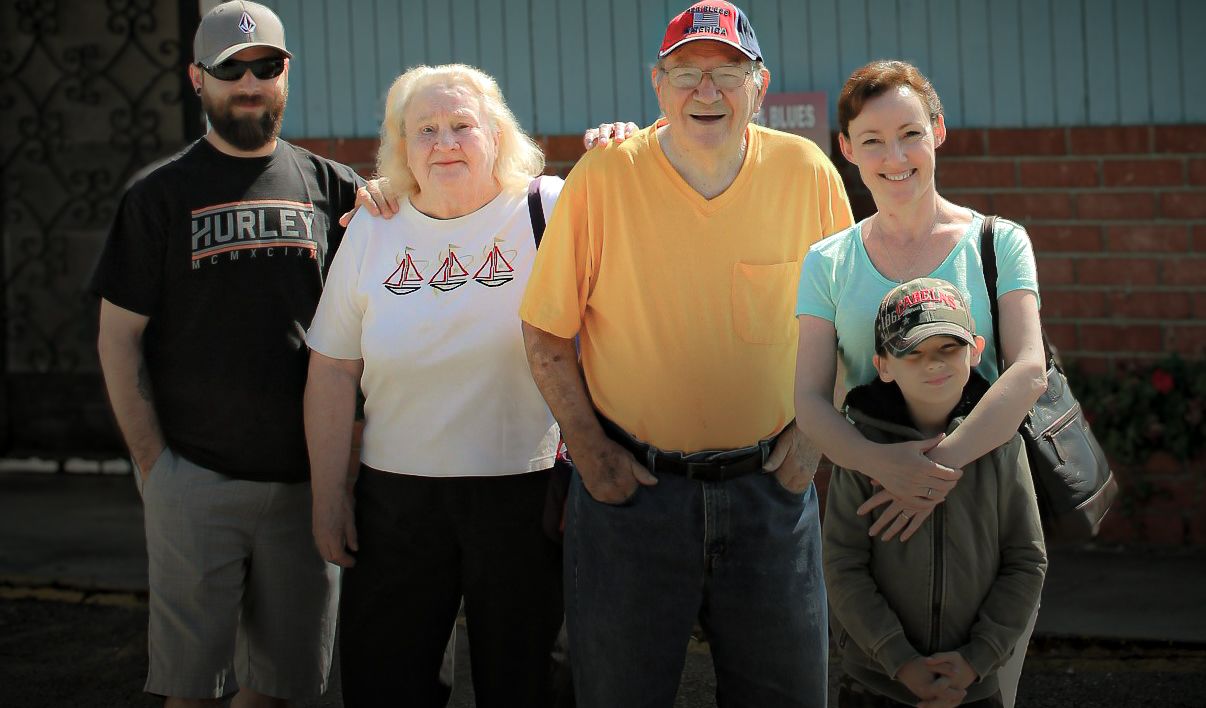 Group of five people posing together outdoors, smiling for a family portrait near a building.