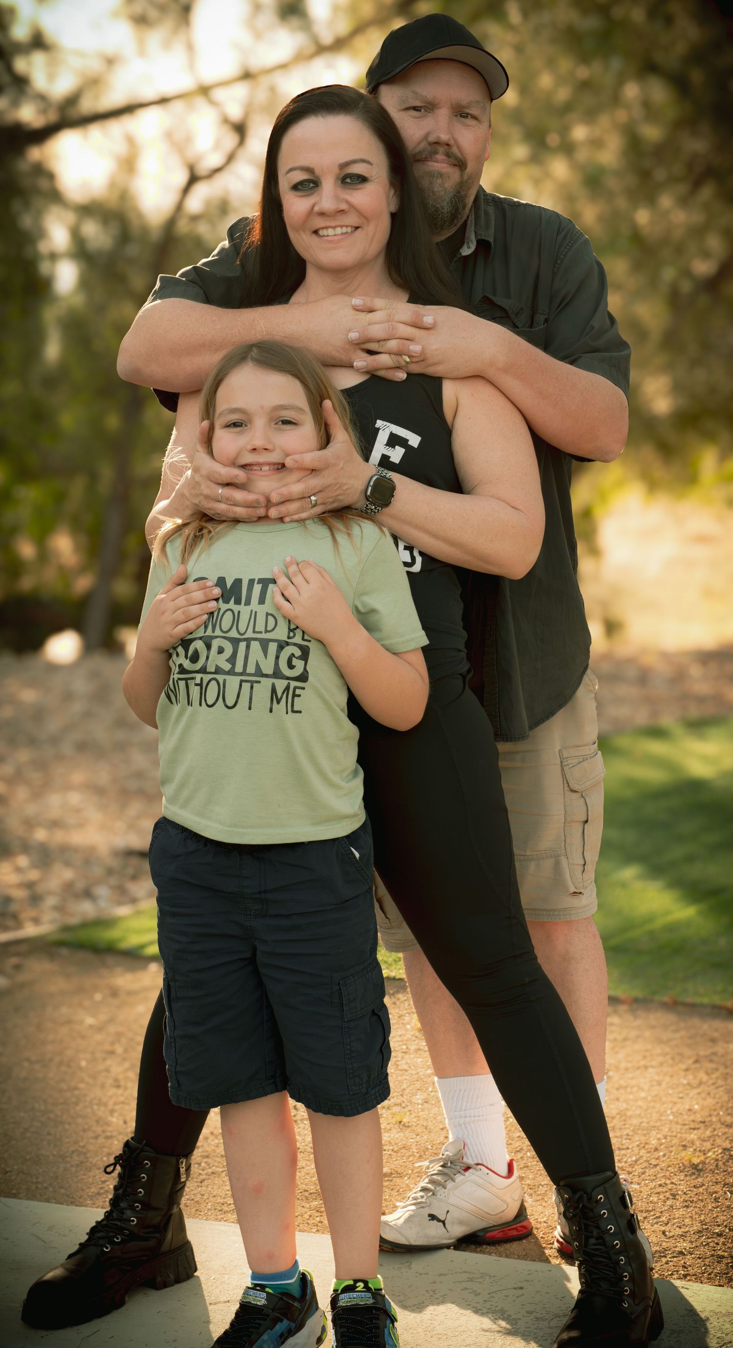 A family of three poses outdoors, with parents hugging their young child in a sunny park setting.