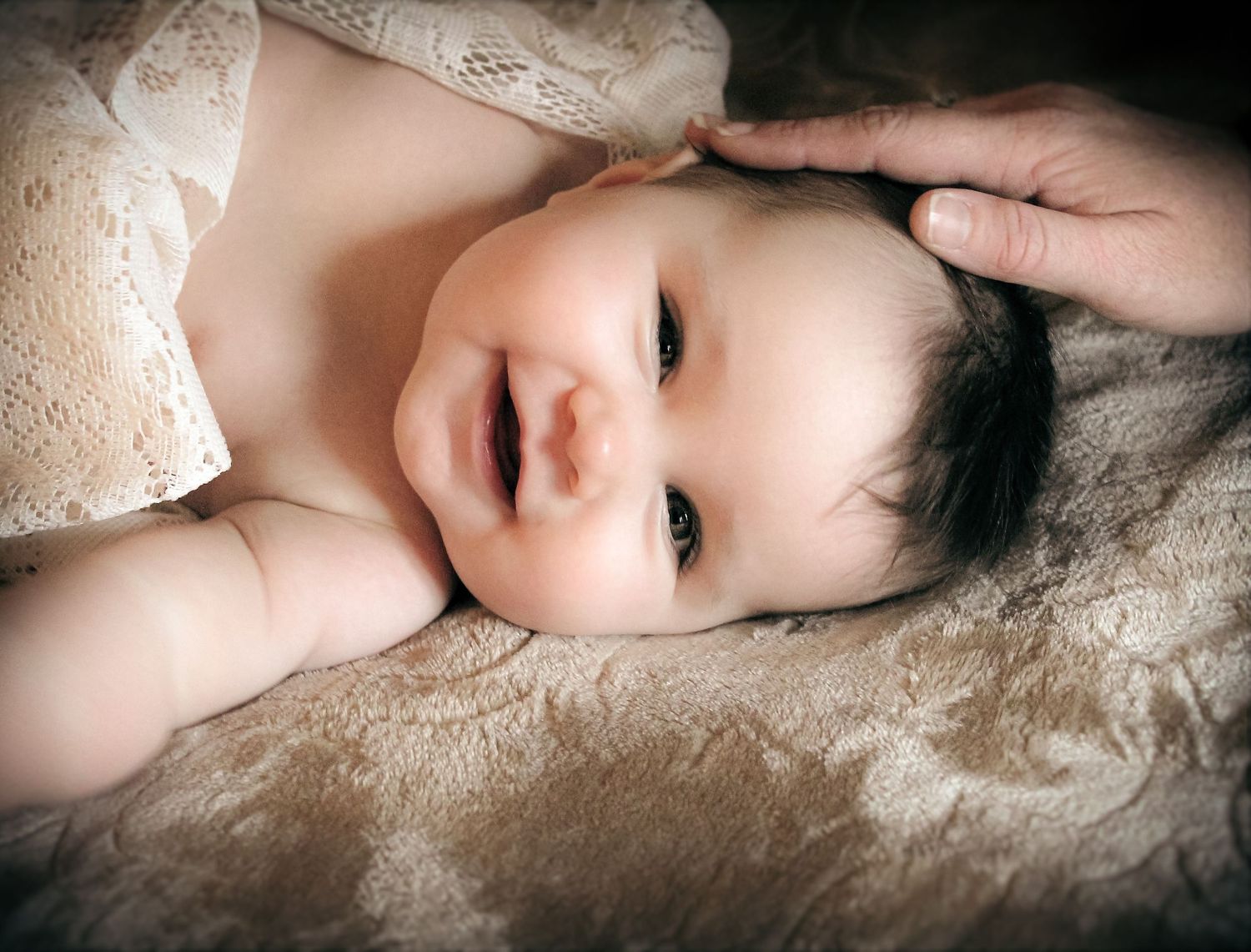 A smiling baby lying on a soft brown blanket with an adult hand gently cradling their head.